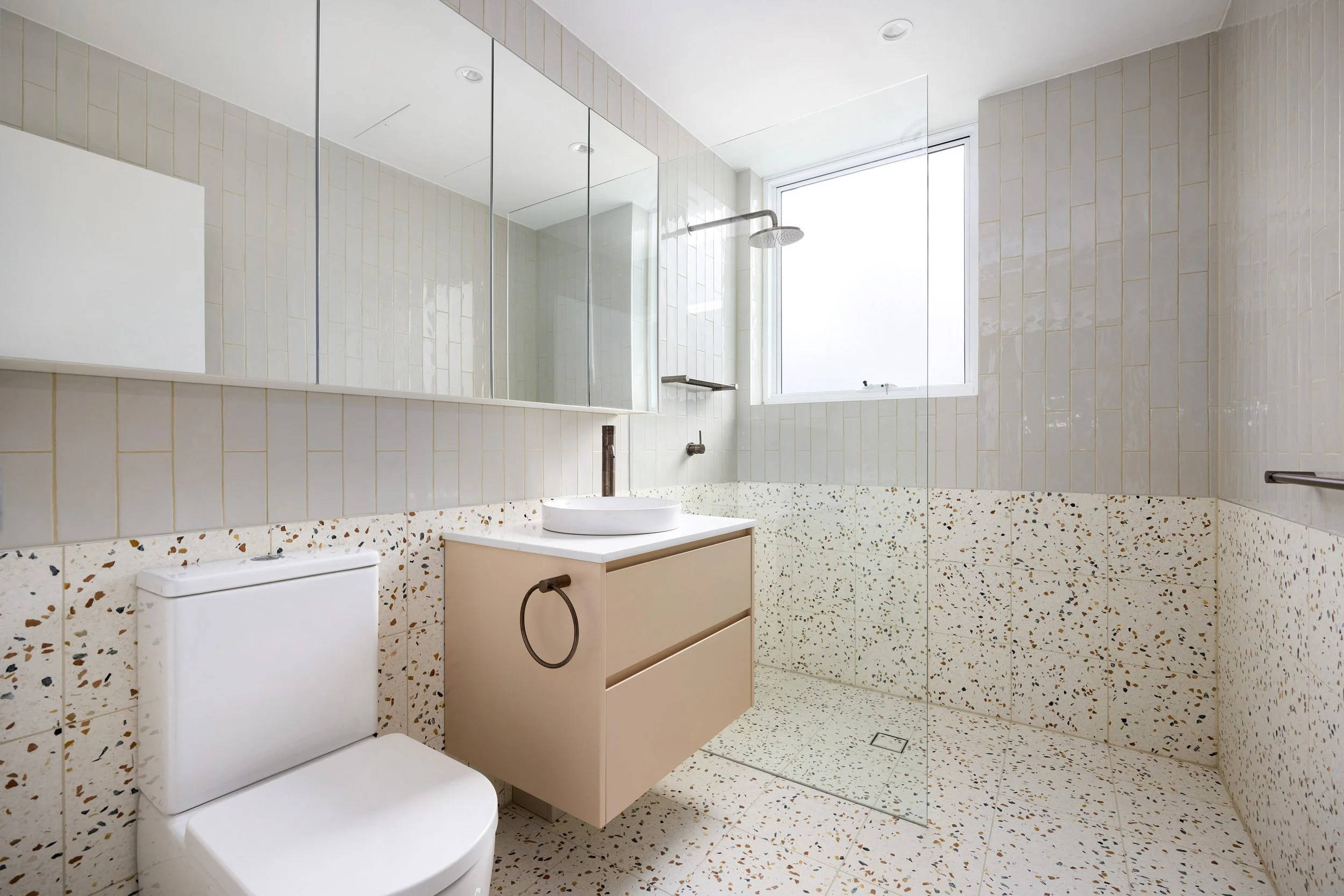 Modern bathroom with a white toilet, beige vanity with a vessel sink, large mirror, terrazzo tiles, glass shower enclosure, and a large window letting in natural light.