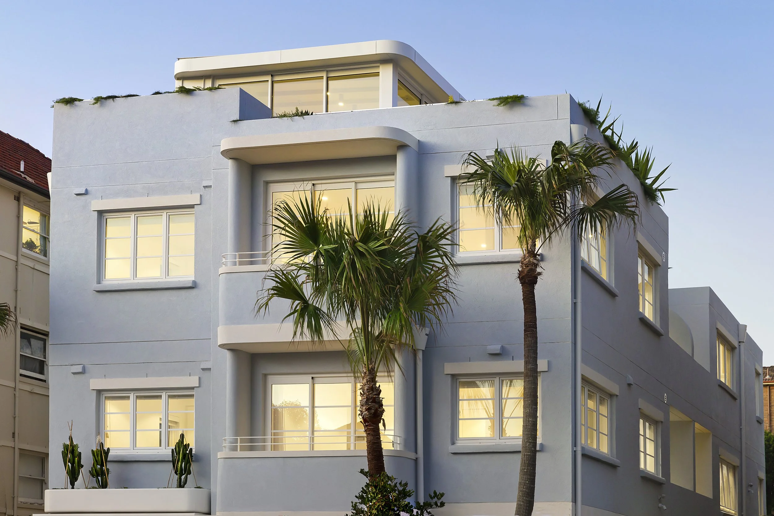 Modern restoration Bondi apartment building with sustainable penthouse extension, large windows, and palm trees in front.