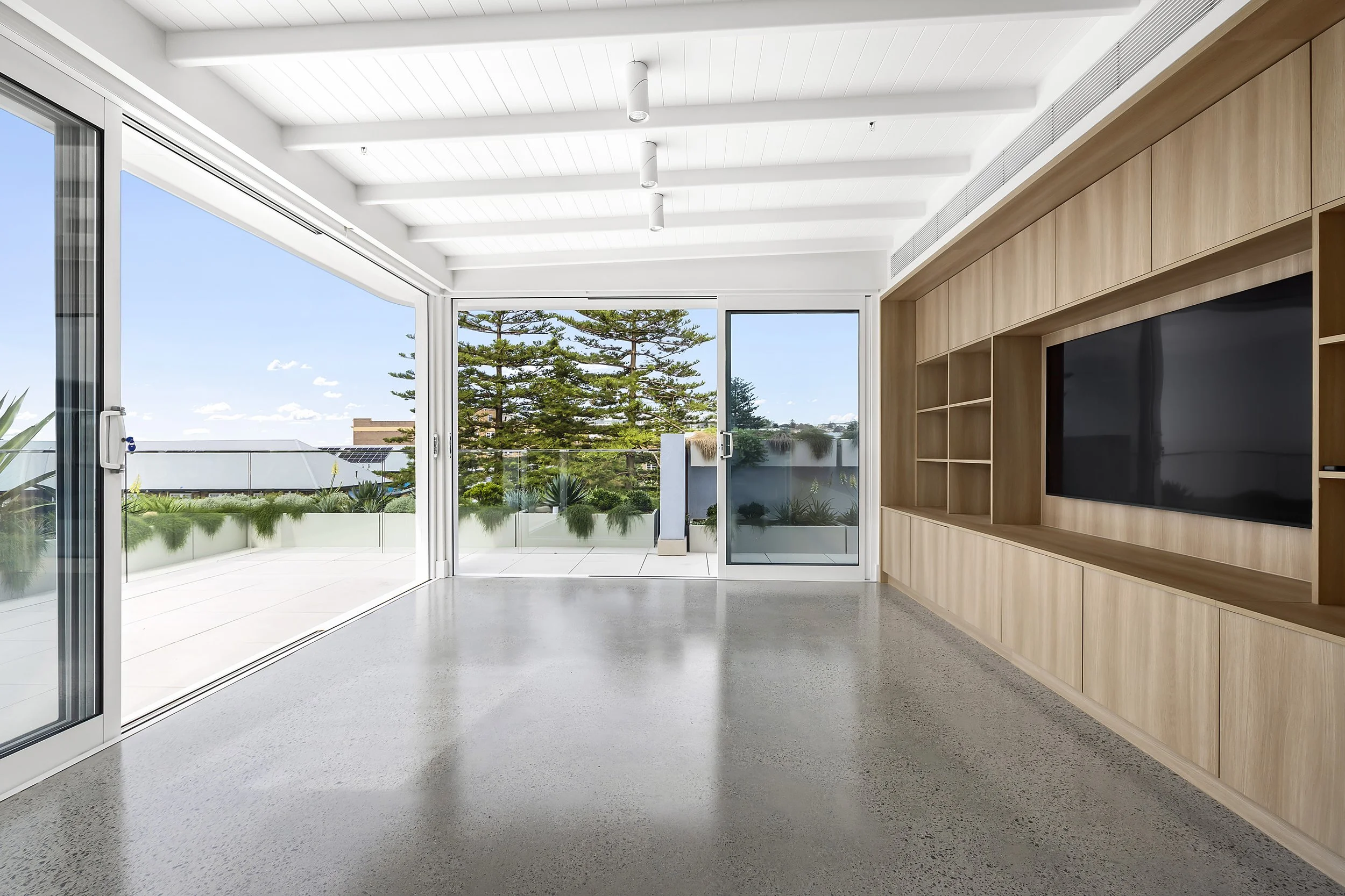 Interior of a modern room with large sliding glass doors opening to a balcony with potted plants and green trees outside. The room has a polished concrete floor, a white ceiling with integrated lighting, and a built-in wooden entertainment unit with 