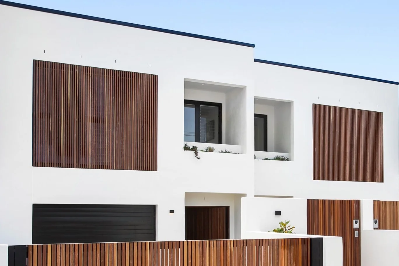 Modern white house with dark wood accents and a flat roof, surrounded by a wooden fence.