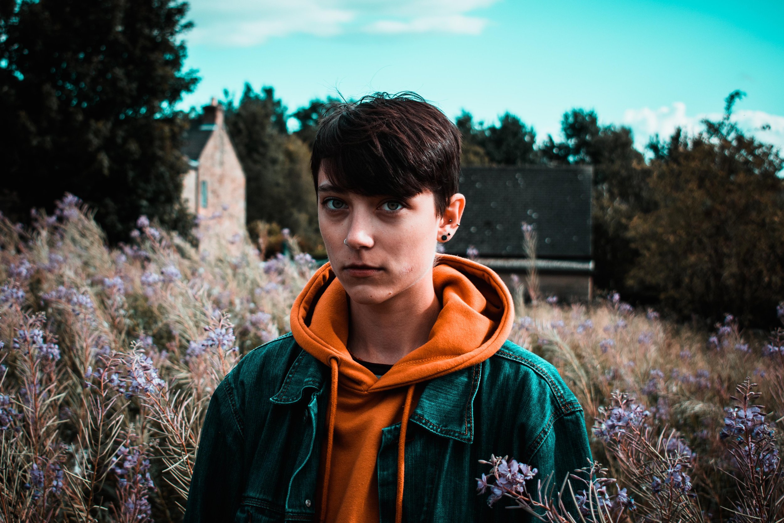 Person with short dark hair wearing an orange hoodie and green jacket standing in a field with purple flowers, trees, and a house in the background.