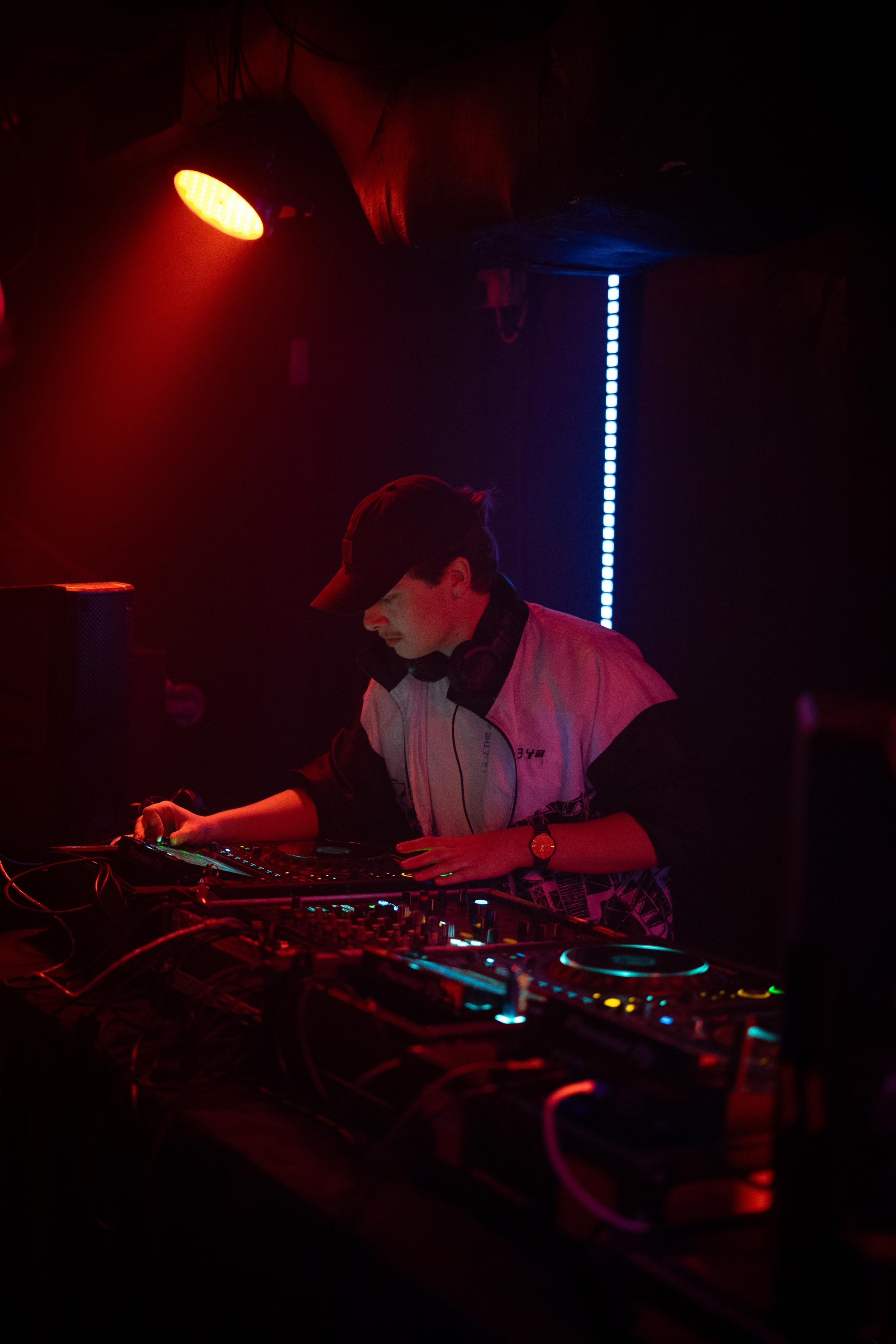DJ performing in dimly lit club with red and blue lights, wearing a cap and headphones around neck, adjusting equipment on a mixer.