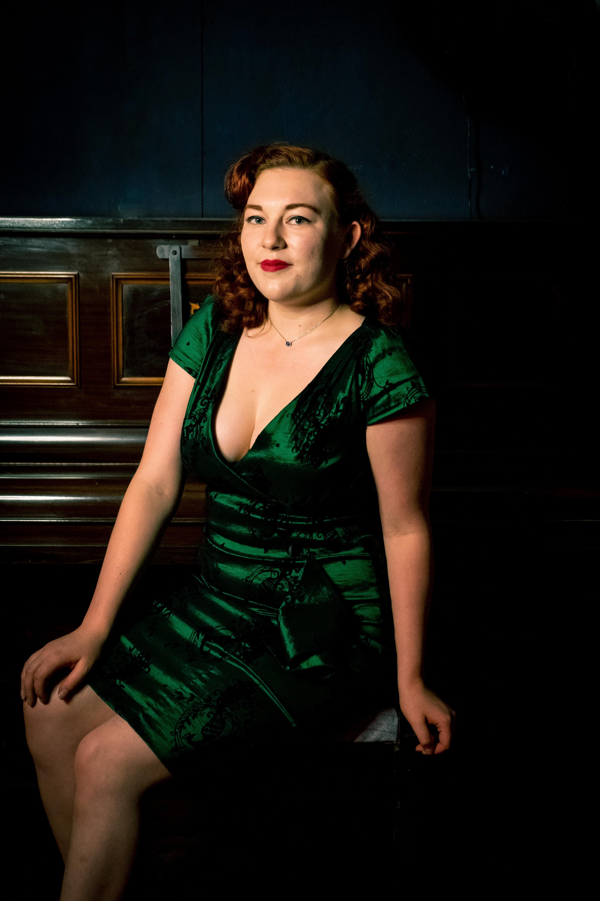 Woman in a green dress sitting on a bench indoors, with dark curly hair and red lipstick.