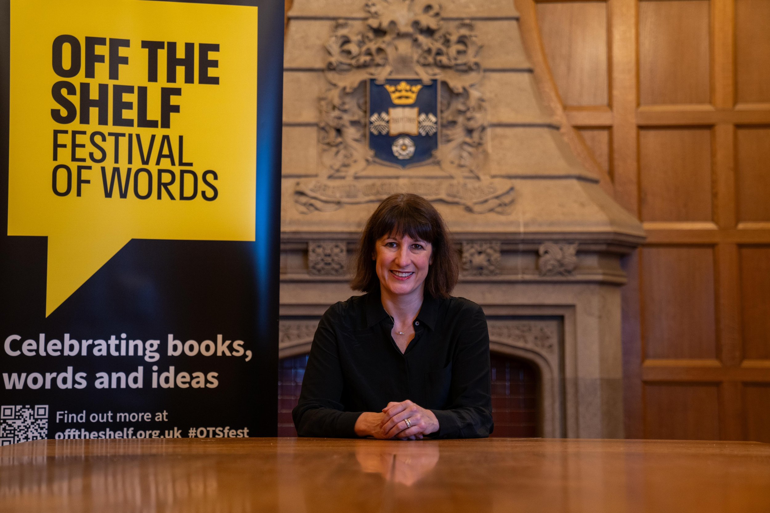 A person seated at a wooden table with a yellow and black banner for 'Off the Shelf Festival of Words' next to them. The background features a decorative fireplace and wood paneling. The banner includes text promoting the festival and a QR code.
