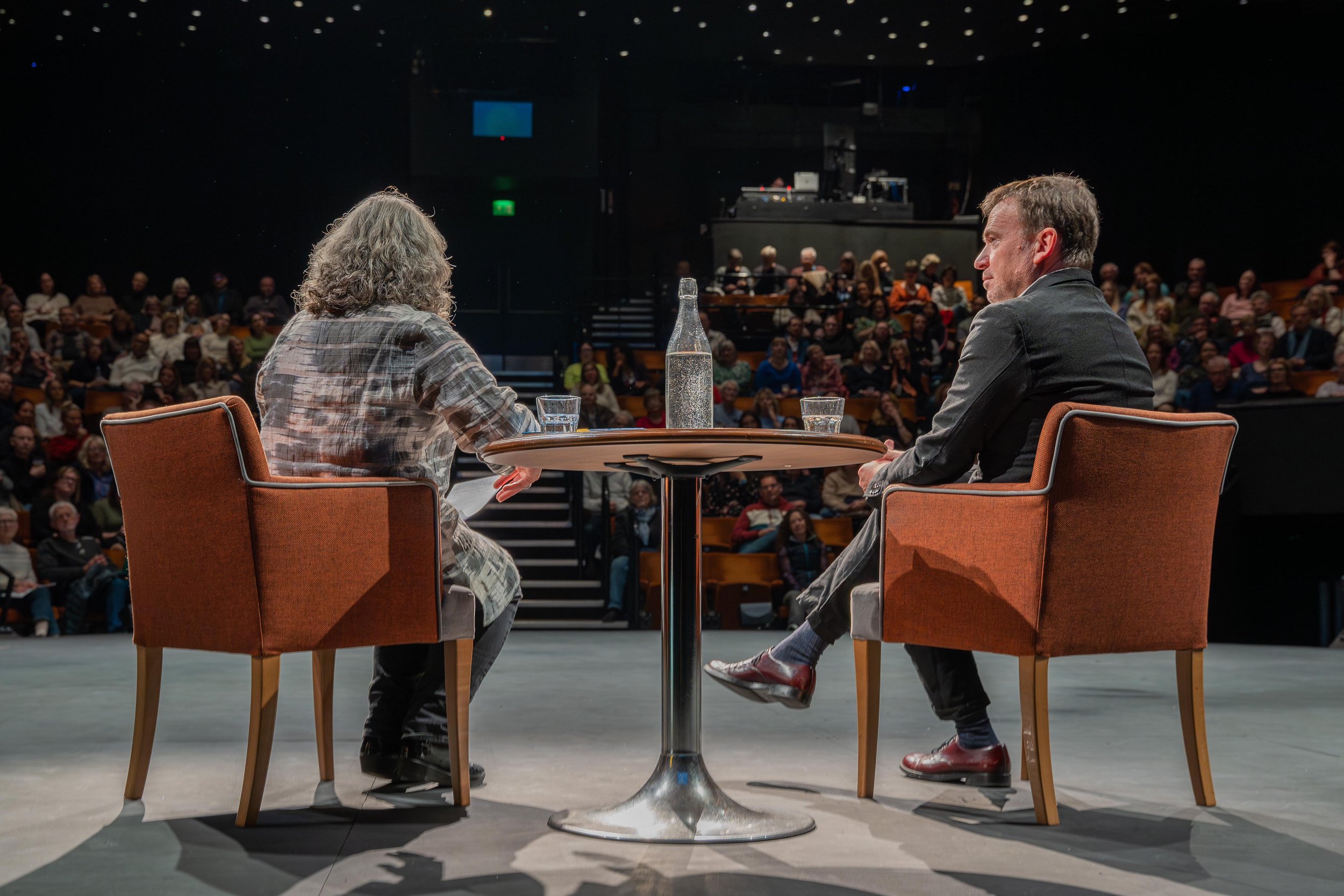 Two people seated on stage, facing an audience, engaged in discussion; a round table with water bottles and glasses between them; audience visible in the background.