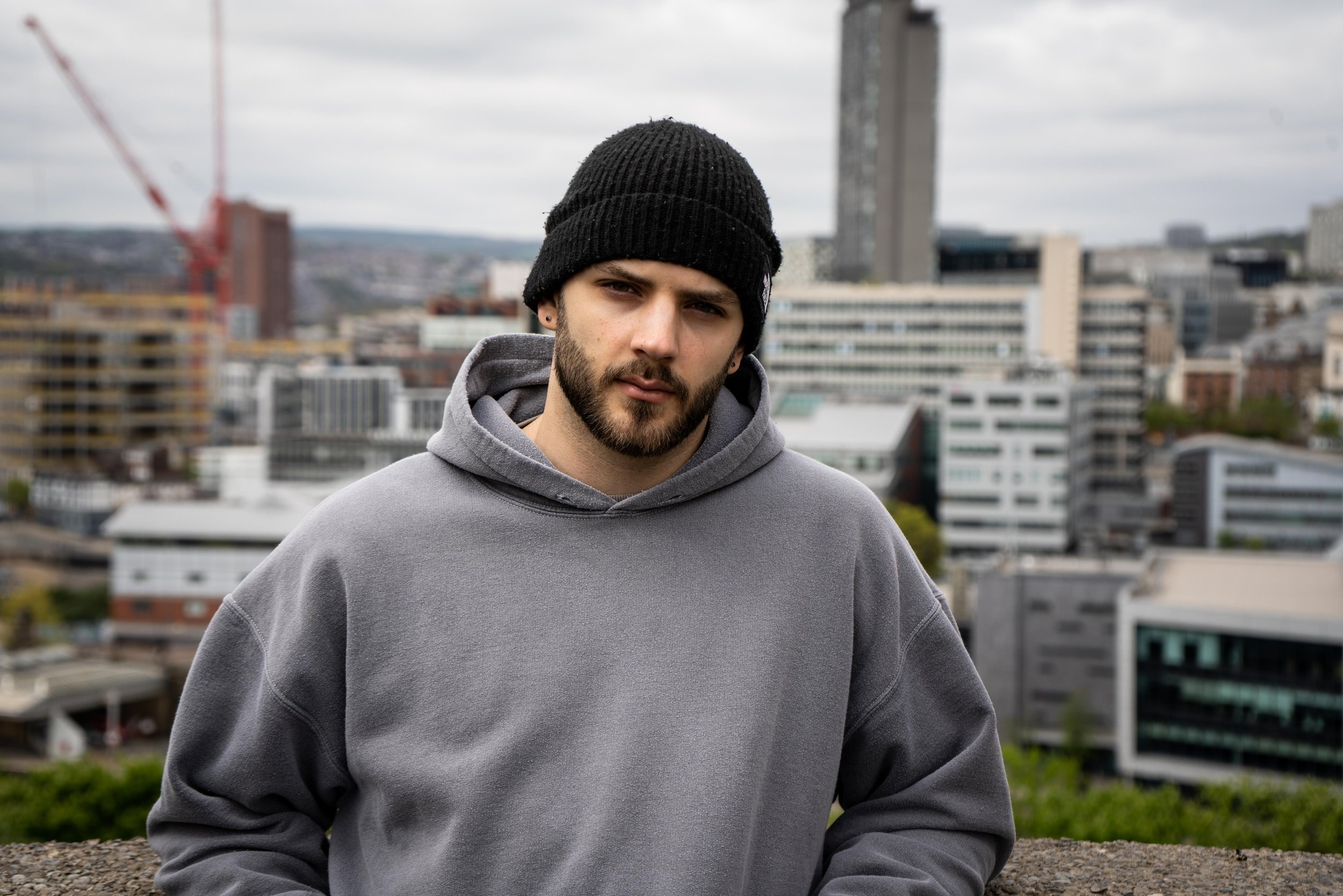 A man in a black beanie and gray hoodie stands in front of a cityscape with modern buildings and a construction crane in the background.