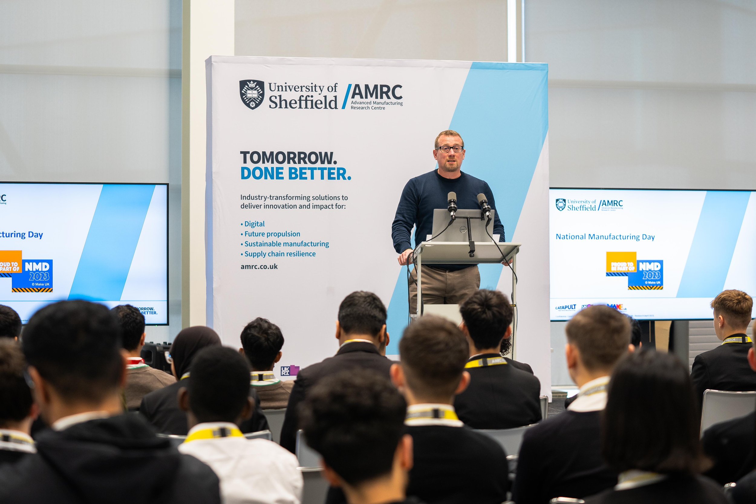 A speaker at a podium with microphones addresses an audience during a University of Sheffield AMRC event. Screens display 'National Manufacturing Day' and related themes. Attendees are seated, facing the speaker.