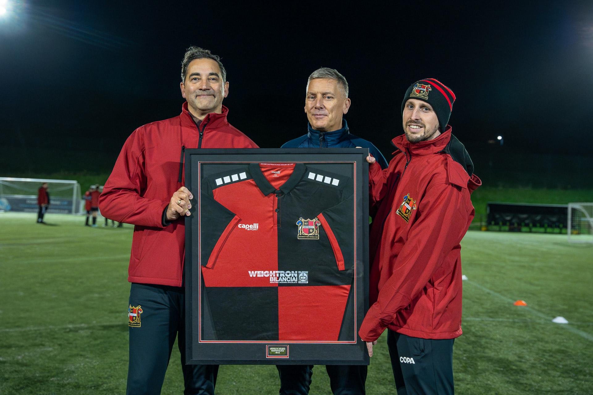 Three people wearing red jackets holding a framed black and red soccer jersey on a lit soccer field at night.