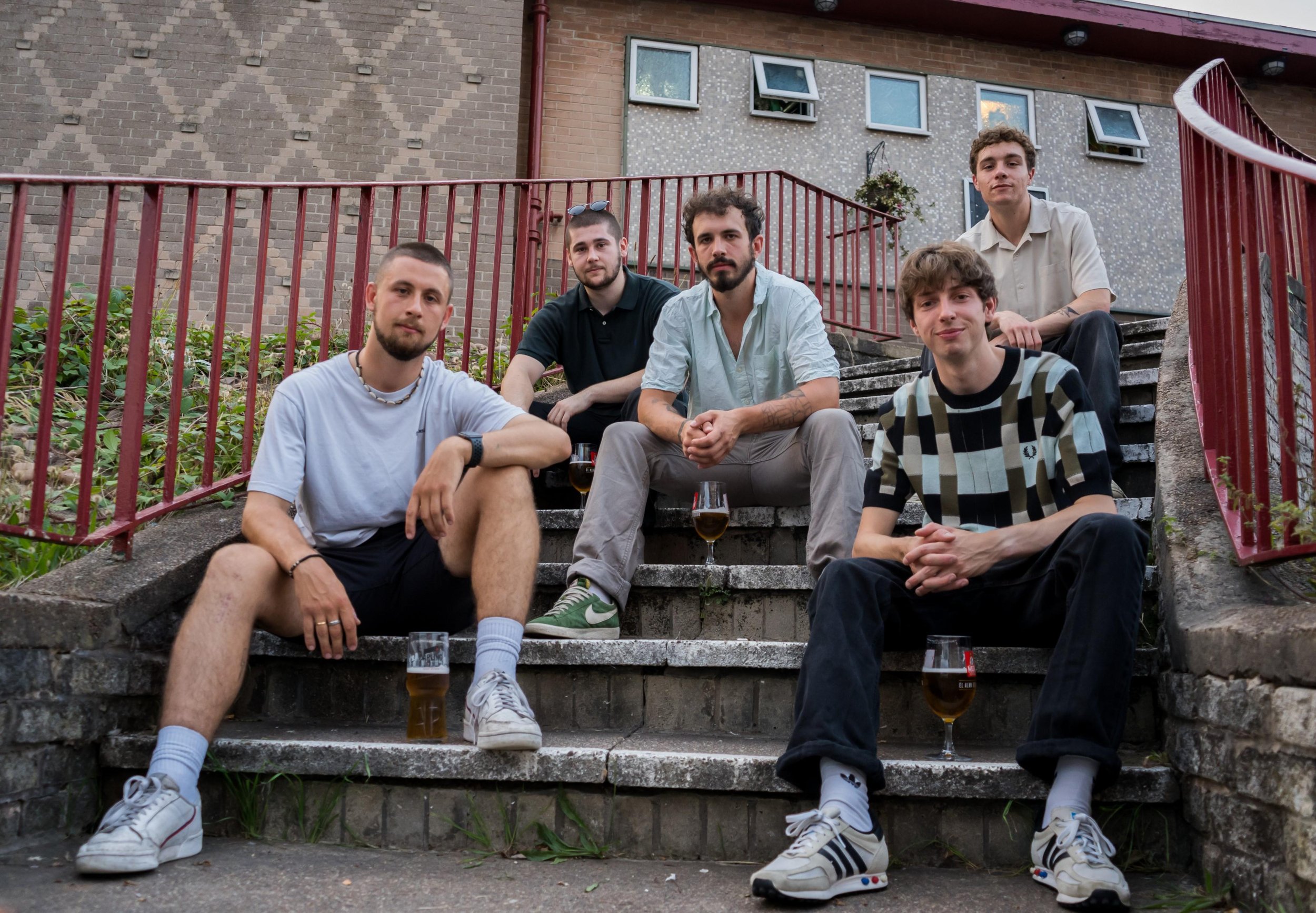 Five young men sitting on outdoor steps with glasses of beer next to them, against a brick building backdrop.
