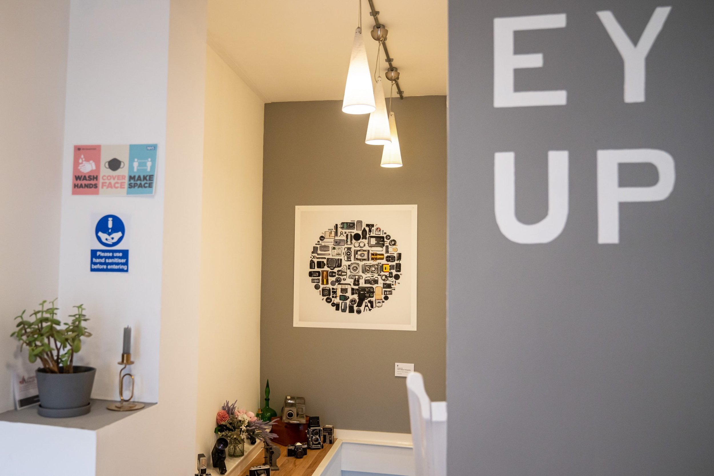 Indoor scene with "EY UP" on a gray wall, a collage of camera illustrations on another wall, pendant lights, a plant, and COVID-19 safety signs.