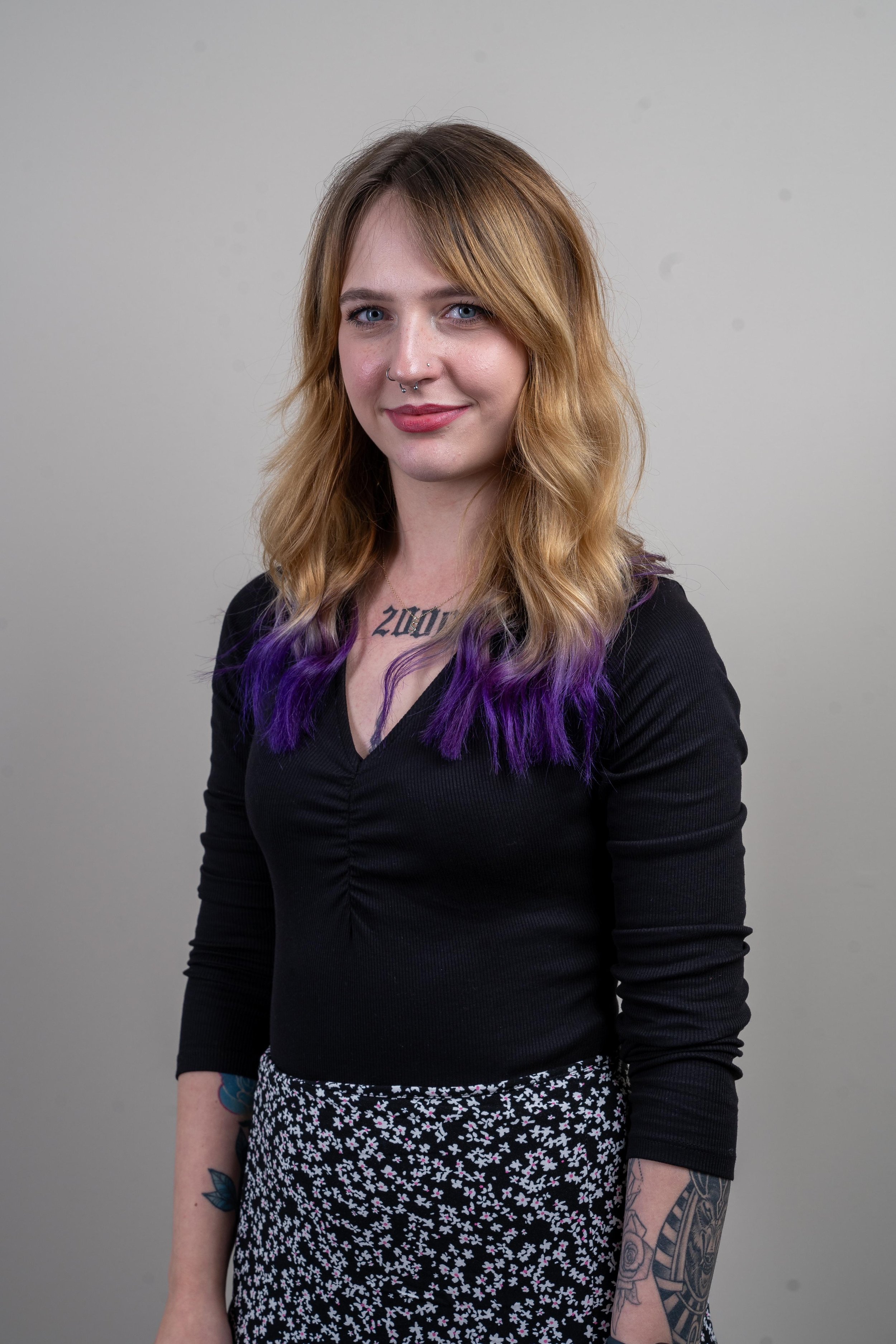 A person with long wavy hair, featuring purple tips, wearing a black top and a floral skirt. Nose and septum pierced, visible tattoo on the forearm.