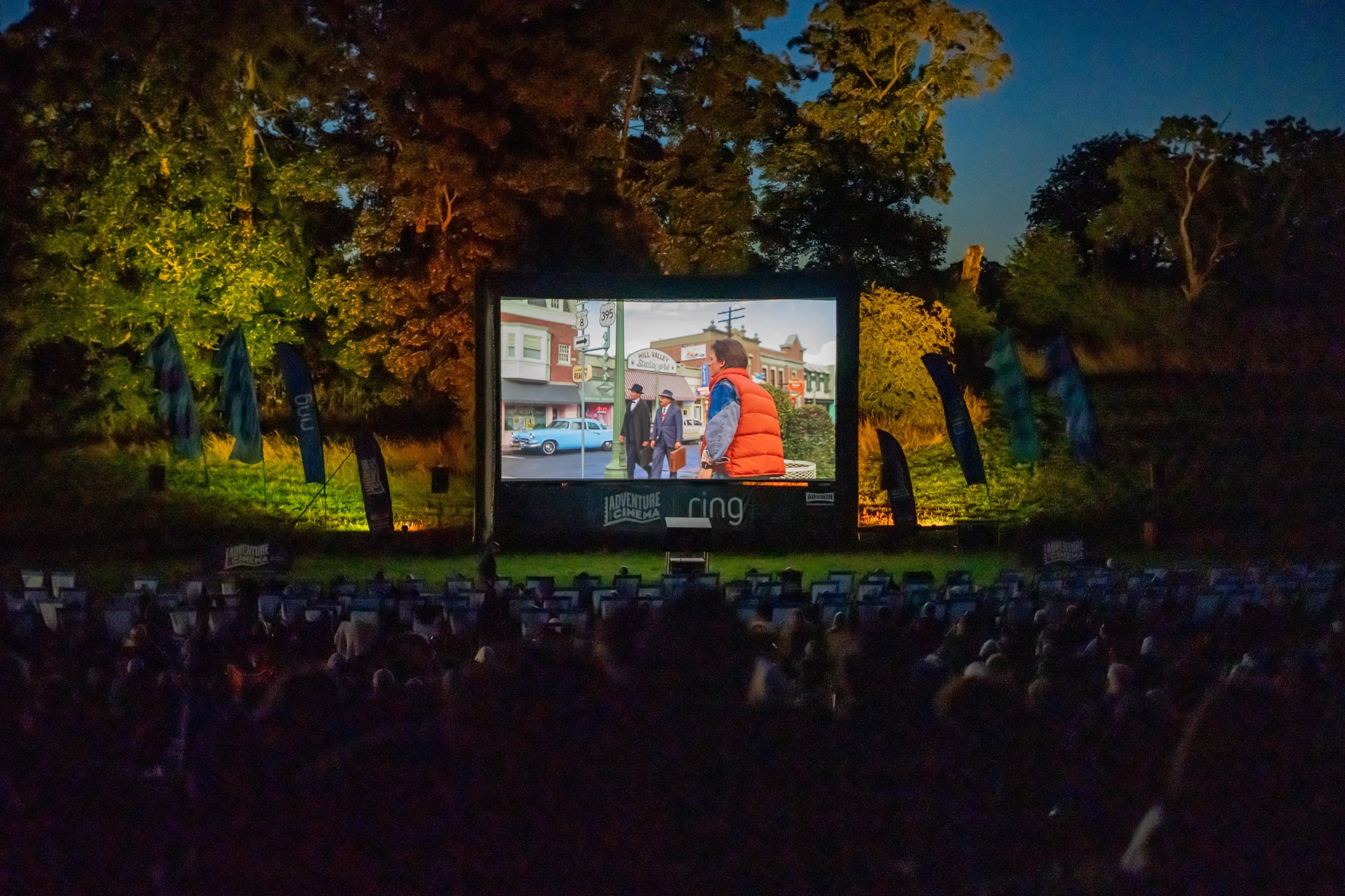 Outdoor cinema screening at night with a crowd watching a movie on a large screen surrounded by trees and illuminated by spotlights.