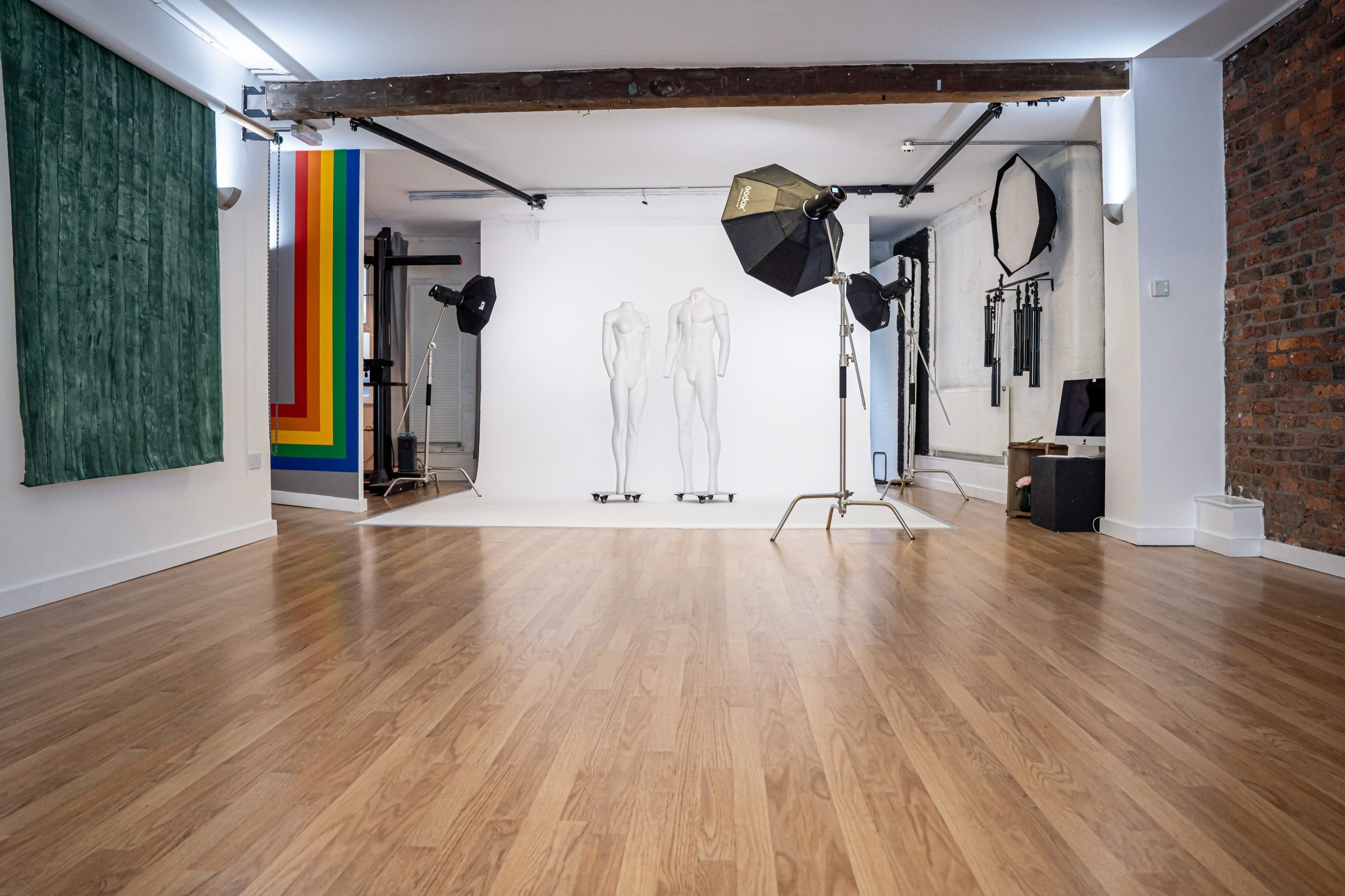 Photography studio with wooden flooring and lighting equipment, including umbrellas and softboxes. Two white mannequins are placed against a white backdrop. A brick wall and colorful striped wall are visible.