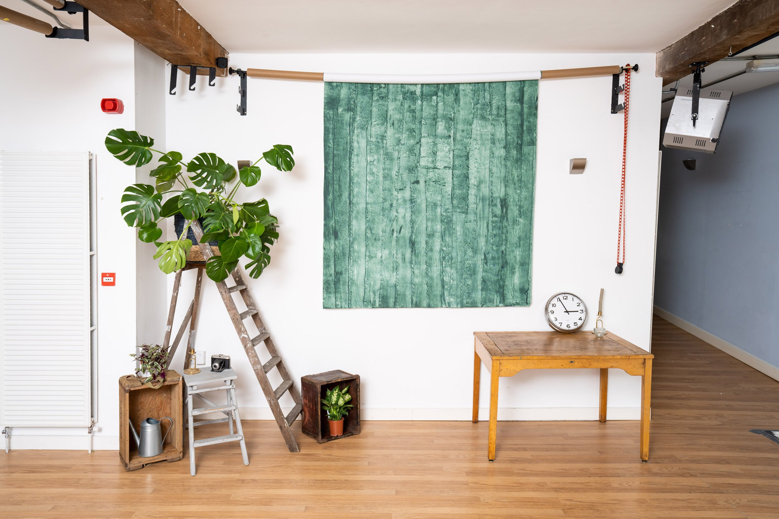 Interior room with wooden ladder, monstera plant, small table, clock, and decorations against white wall and green fabric backdrop.