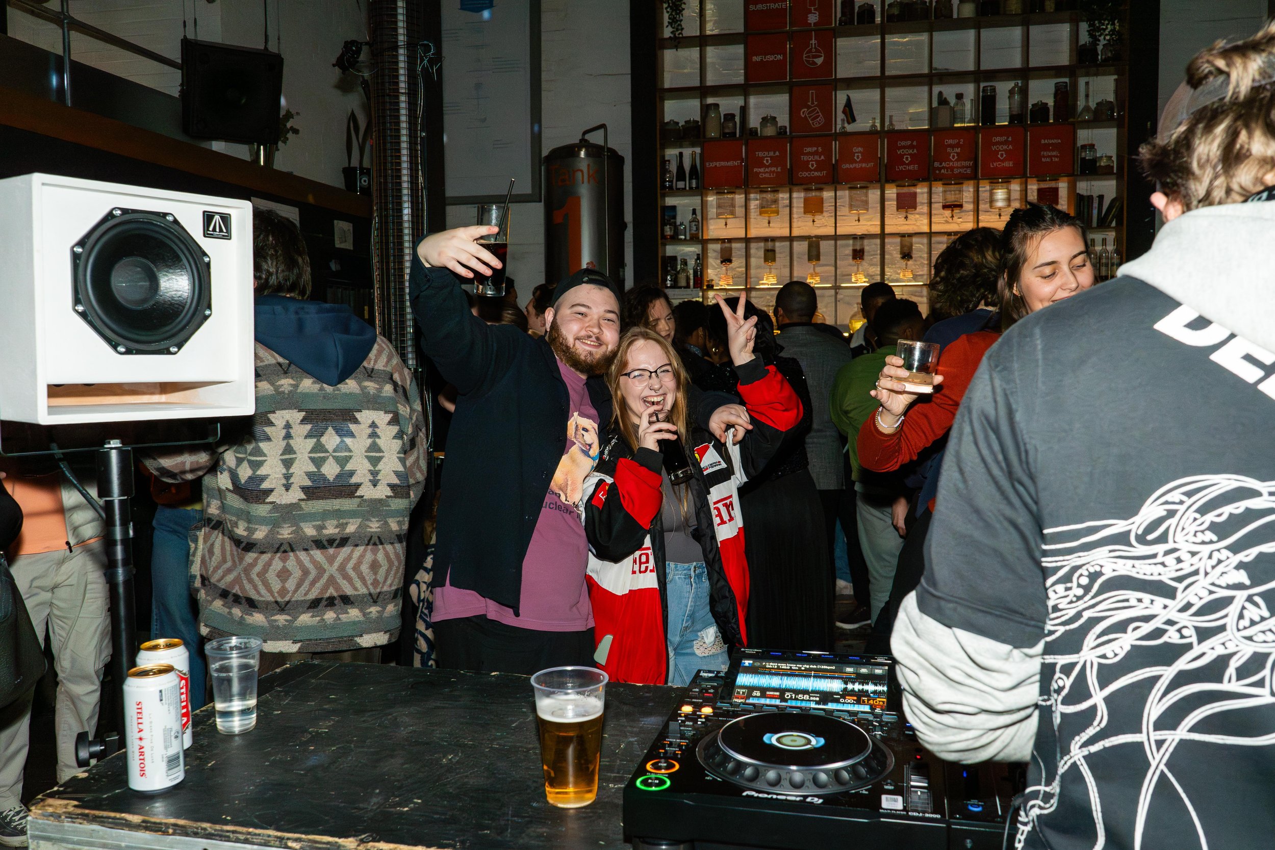People enjoying a party, smiling and posing for the camera, with a DJ setup and drinks.