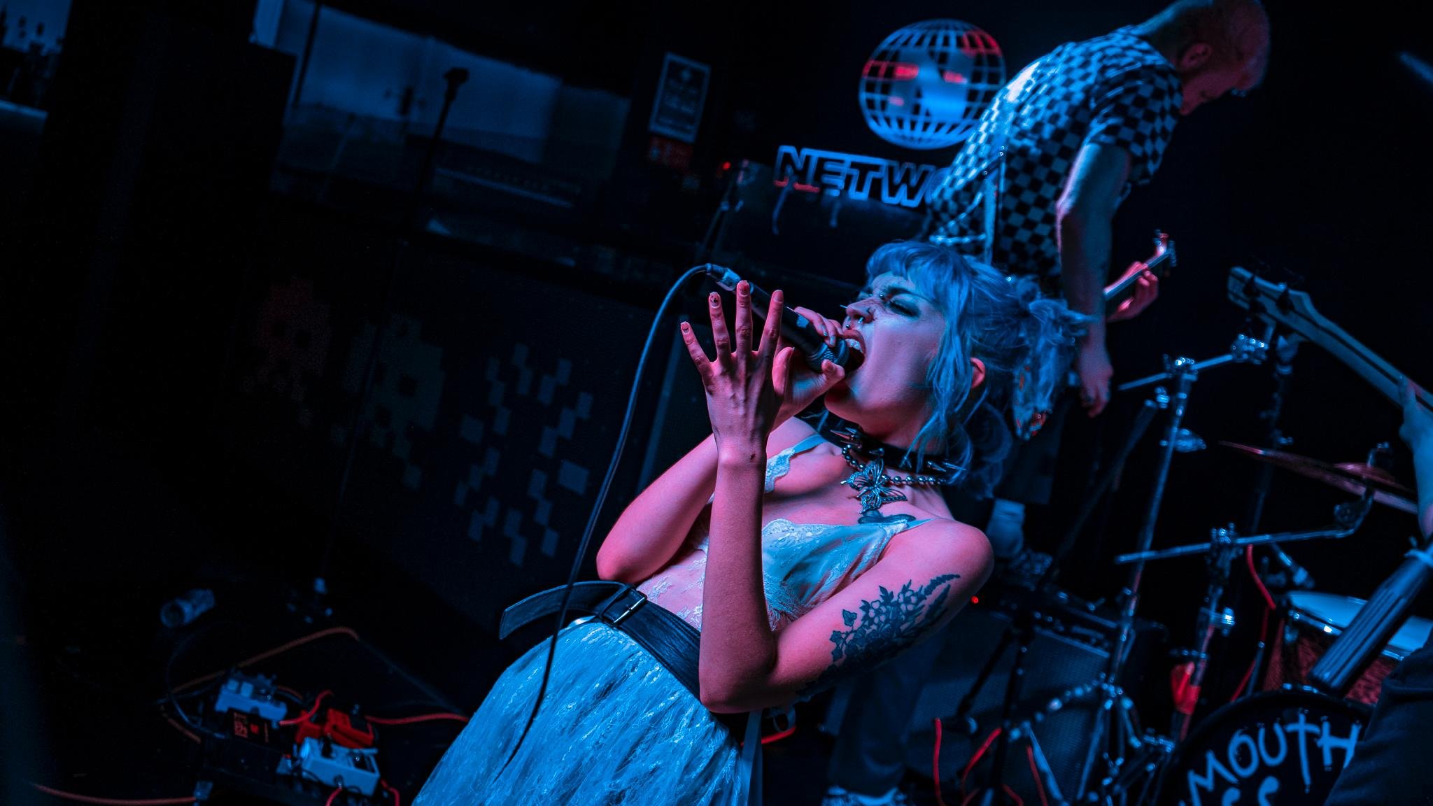 Band performing live on stage; female vocalist singing passionately into a microphone, wearing a choker and a blue dress, tattoos on her arm; drummer playing in the background; dark ambient lighting.