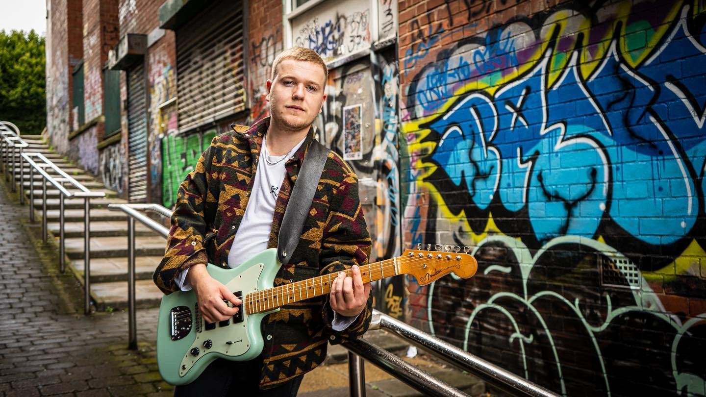 Man playing an electric guitar in front of graffiti-covered wall on urban street with stairs and metal railing.