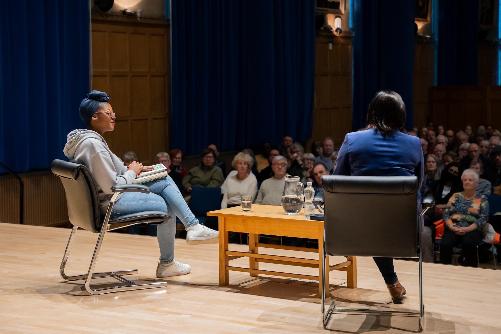 Two people seated on stage with an audience in the background, engaged in a discussion. One person is wearing a gray hoodie and blue jeans, holding papers, while the other is in a blue suit. A wooden table with water pitchers and glasses is between t