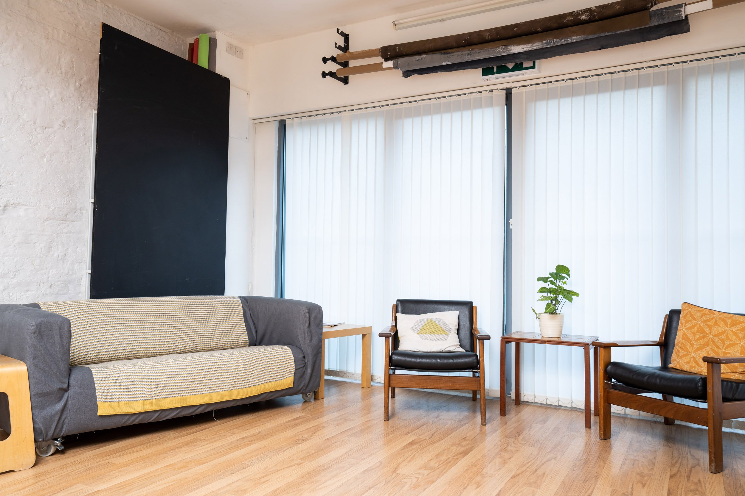 Modern living room with gray sofa, two wooden armchairs, light wood flooring, vertical blinds, and a potted plant on a side table.