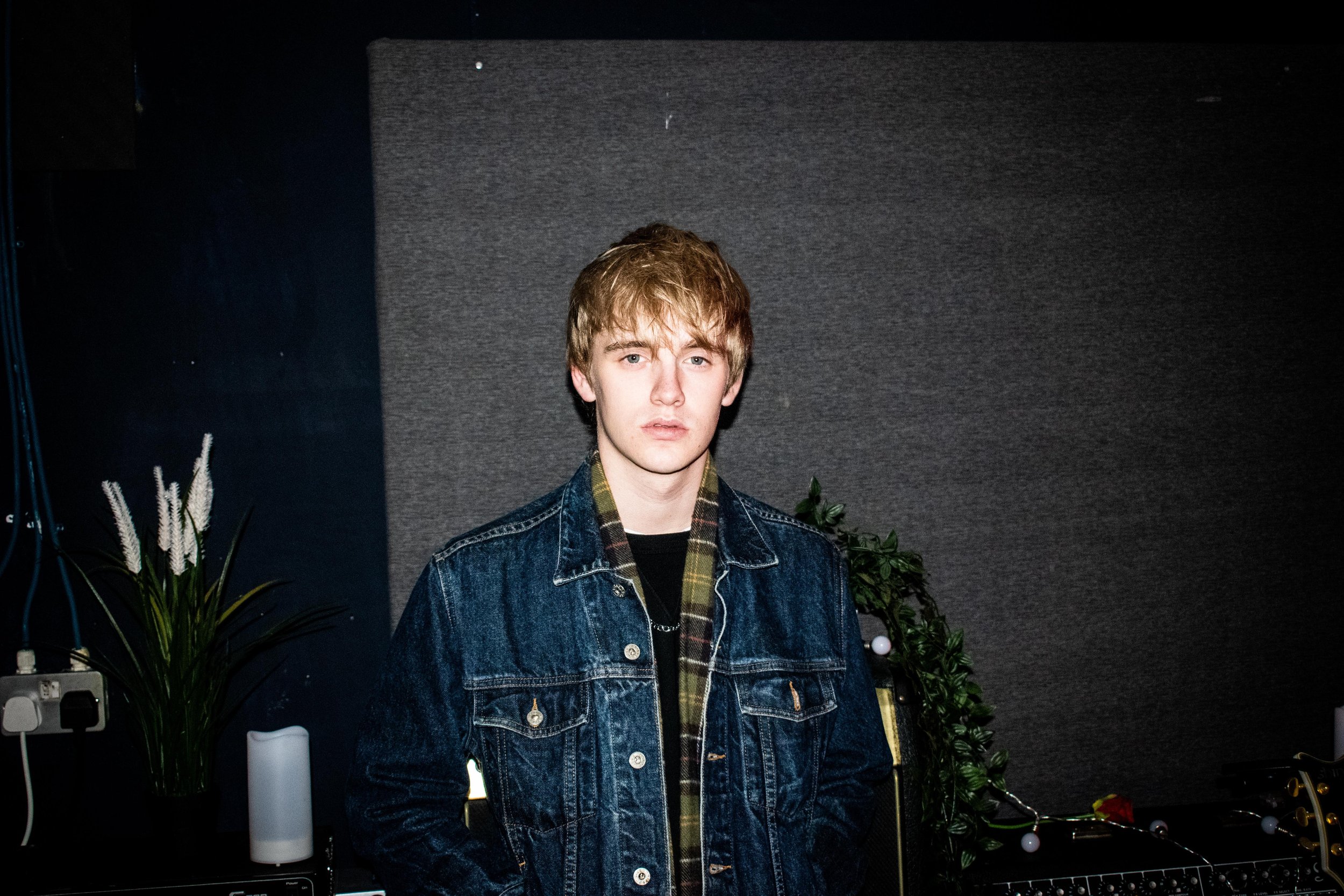Person wearing a denim jacket, standing indoors, with a plant and audio equipment in the background.