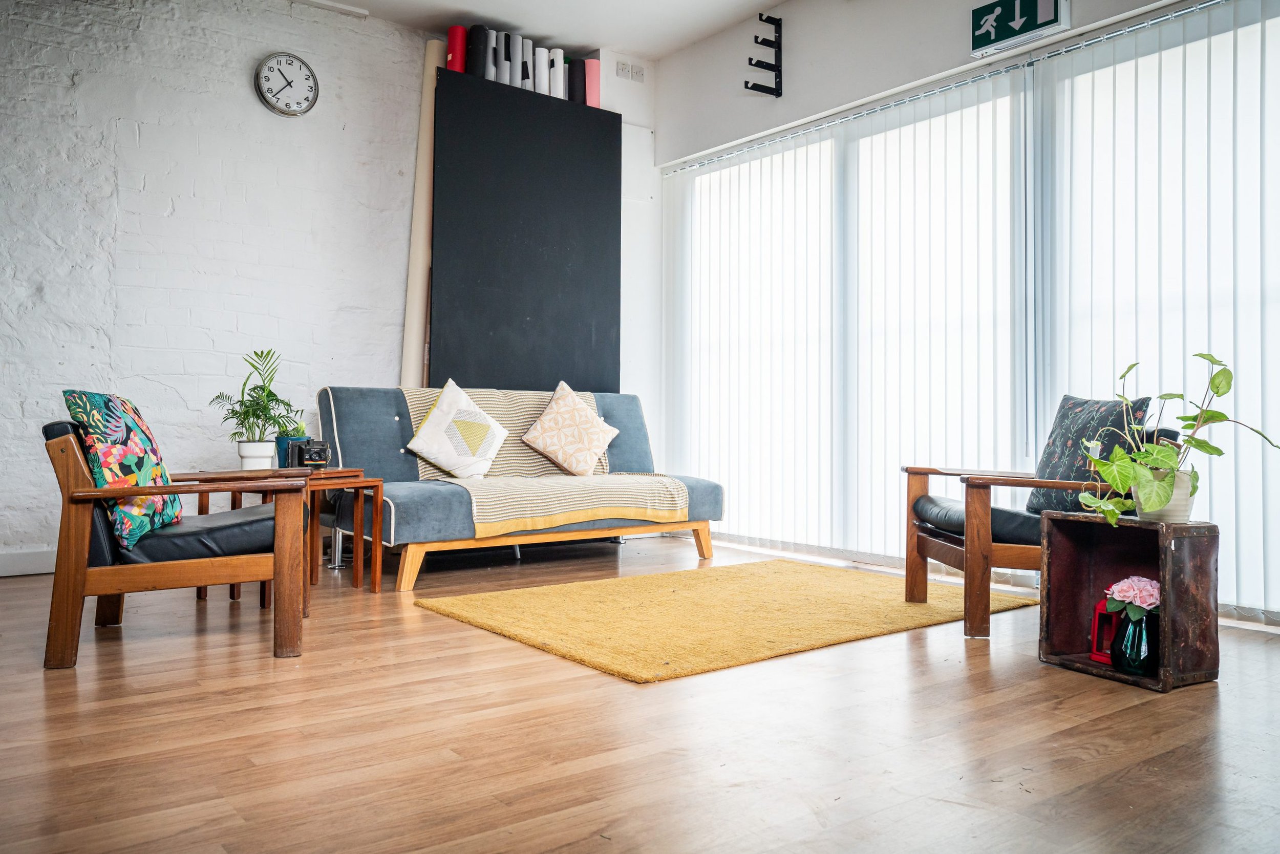 A modern living room with a minimalist design, featuring wooden furniture, a blue sofa with decorative cushions, a yellow area rug, and a wooden floor. The room includes large windows with vertical blinds, a wall clock, potted plants, and a small tab