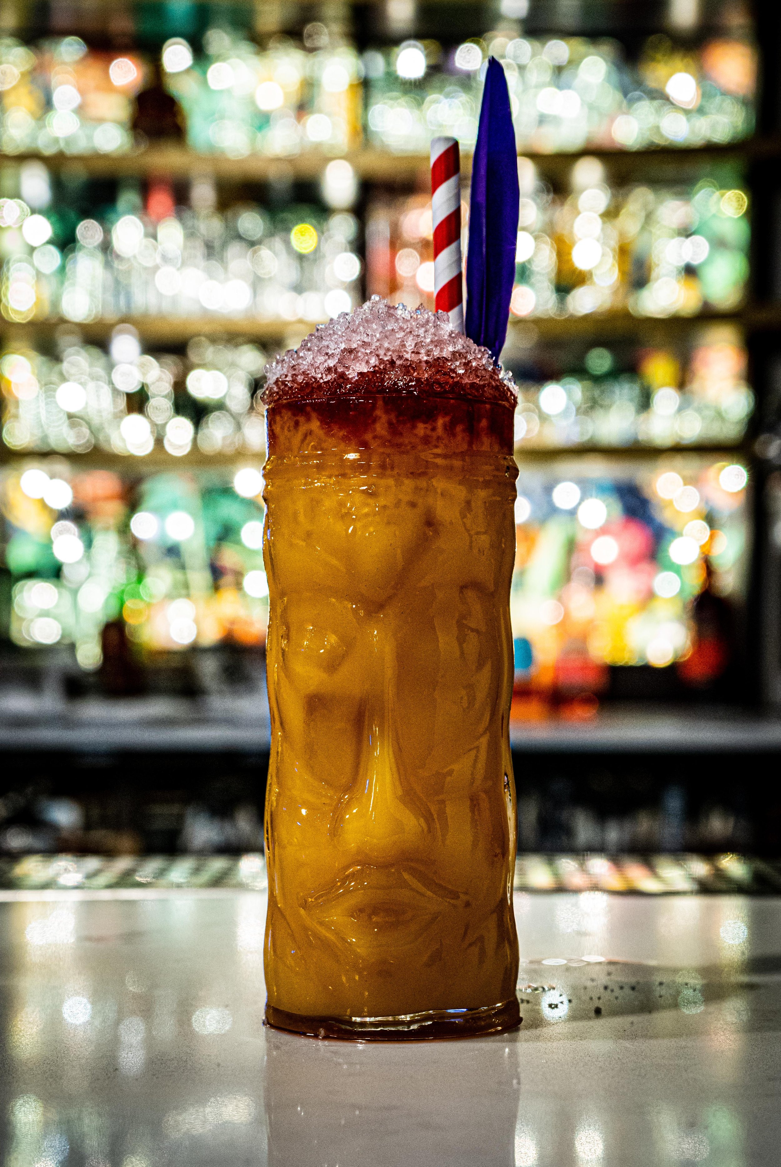 Tiki-style cocktail glass with a colorful drink topped with crushed ice and garnished with a straw and feather, set against a blurred bar background.