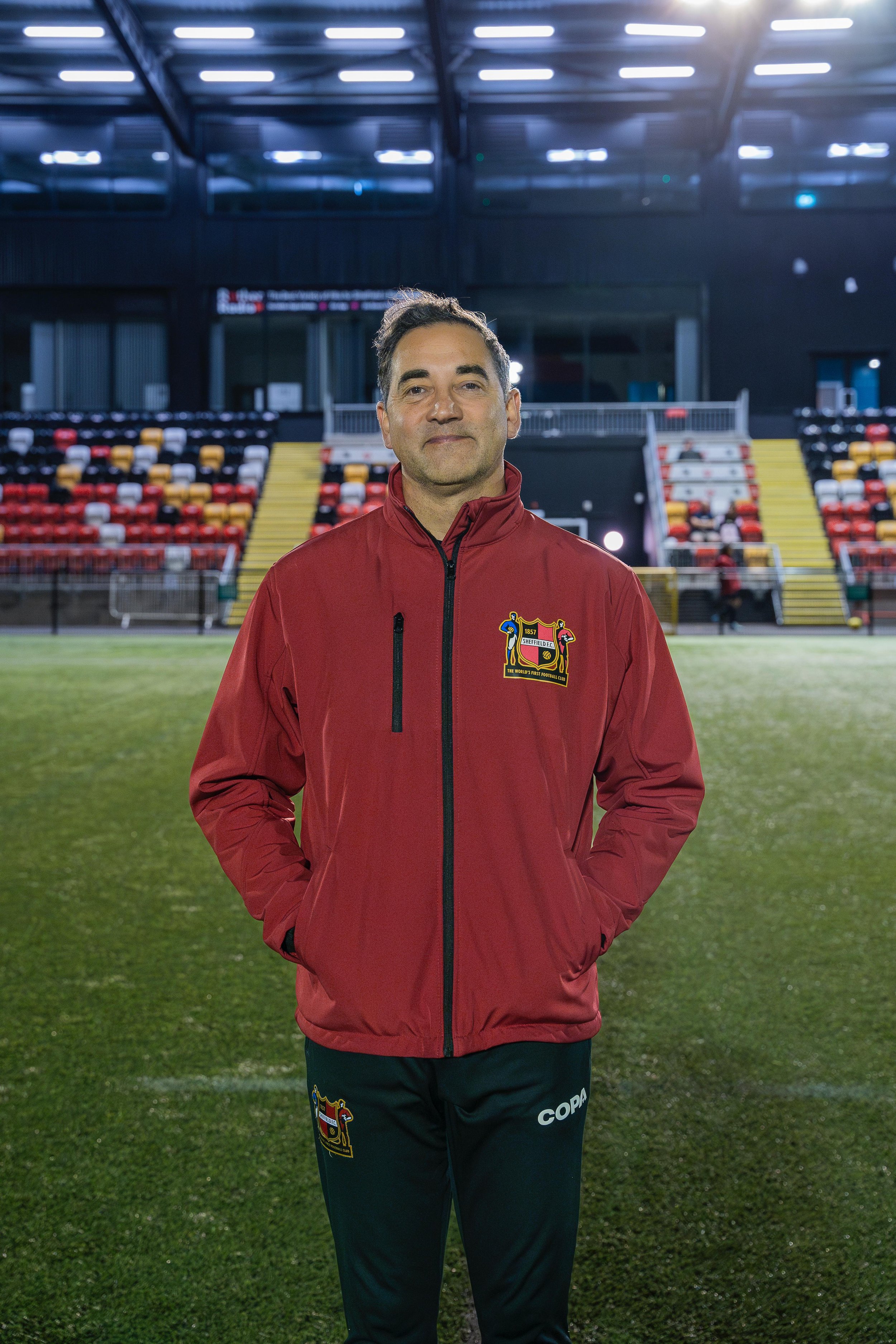 A person standing on a soccer field wearing a red jacket with hands in pockets; stadium seats in the background.
