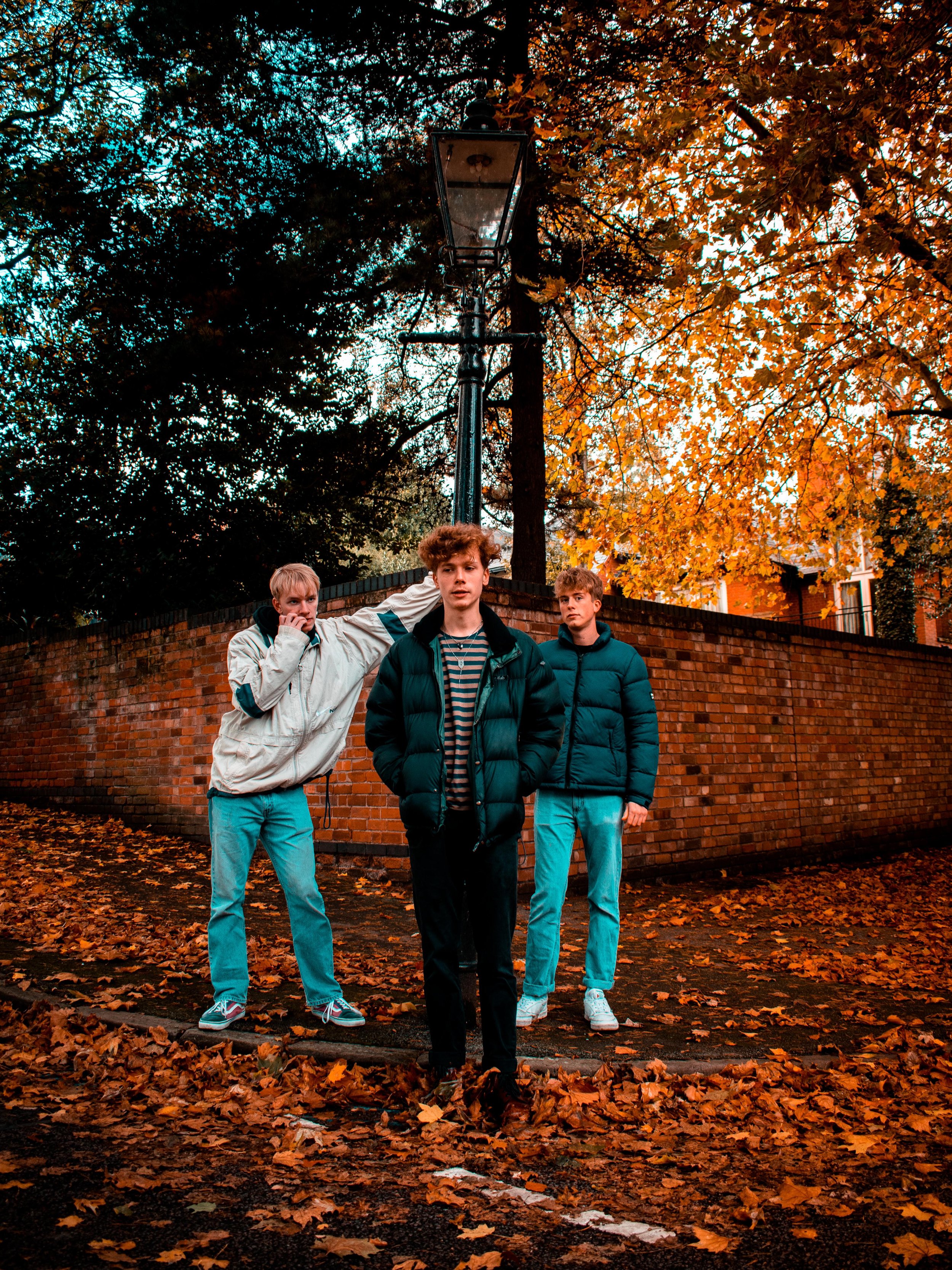 Three young men standing outdoors on a leaf-covered ground, wearing casual jackets and jeans, with autumn trees and a brick wall in the background. A streetlamp is positioned behind them.