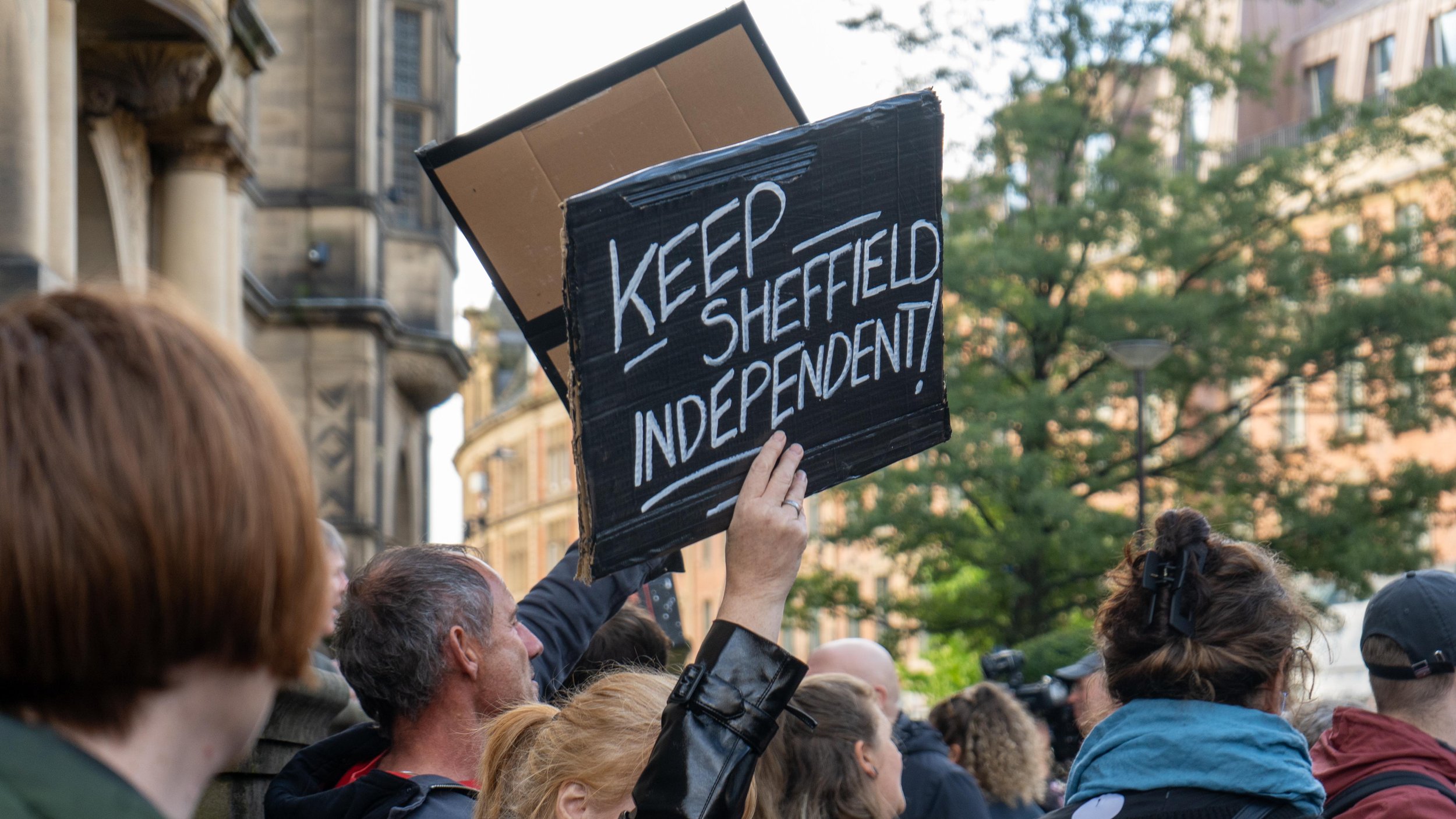 People holding a "Keep Sheffield Independent!" sign during a protest or demonstration in an urban area.