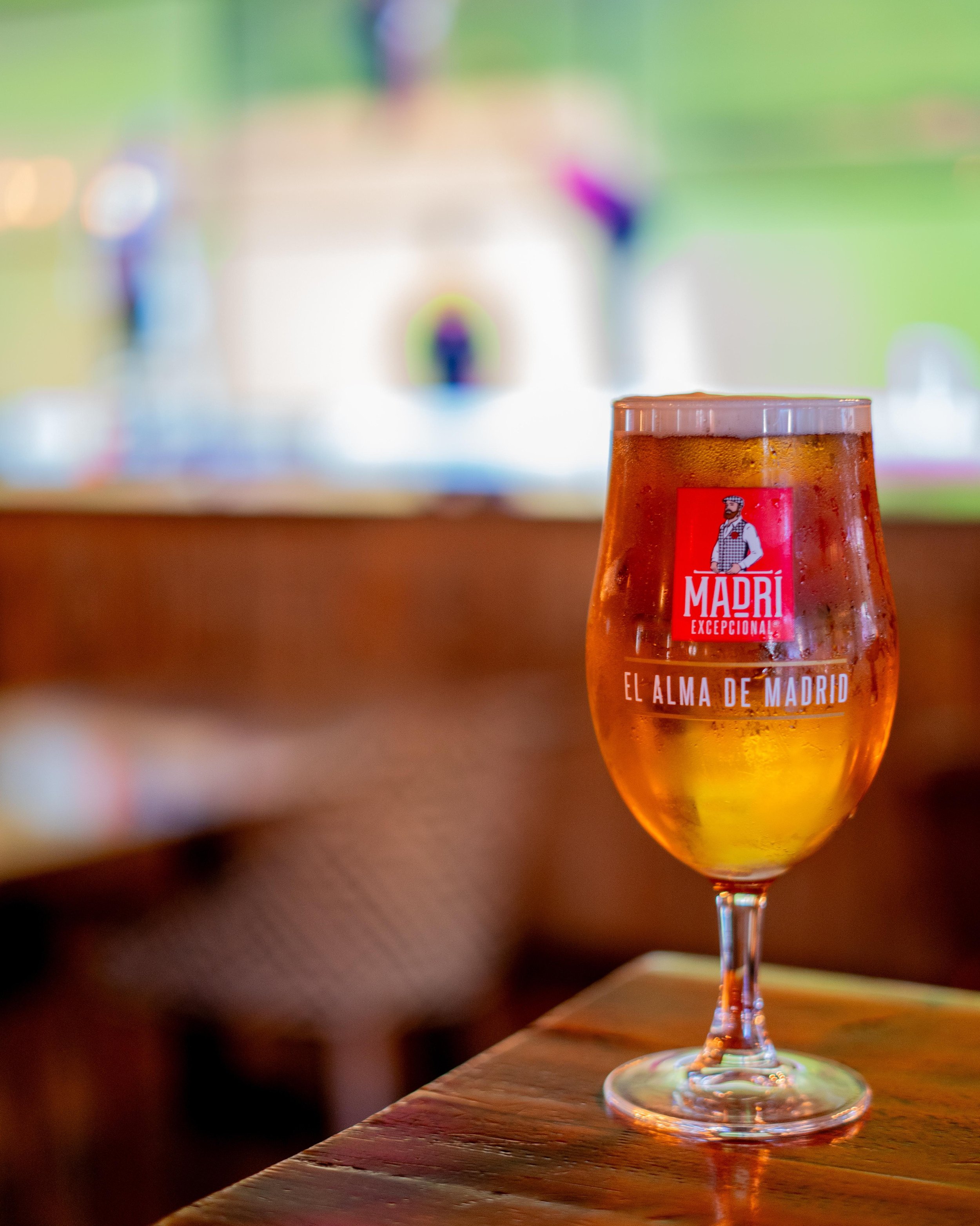 Glass of beer labeled "Madri Excepcional" on a wooden table with blurred background.