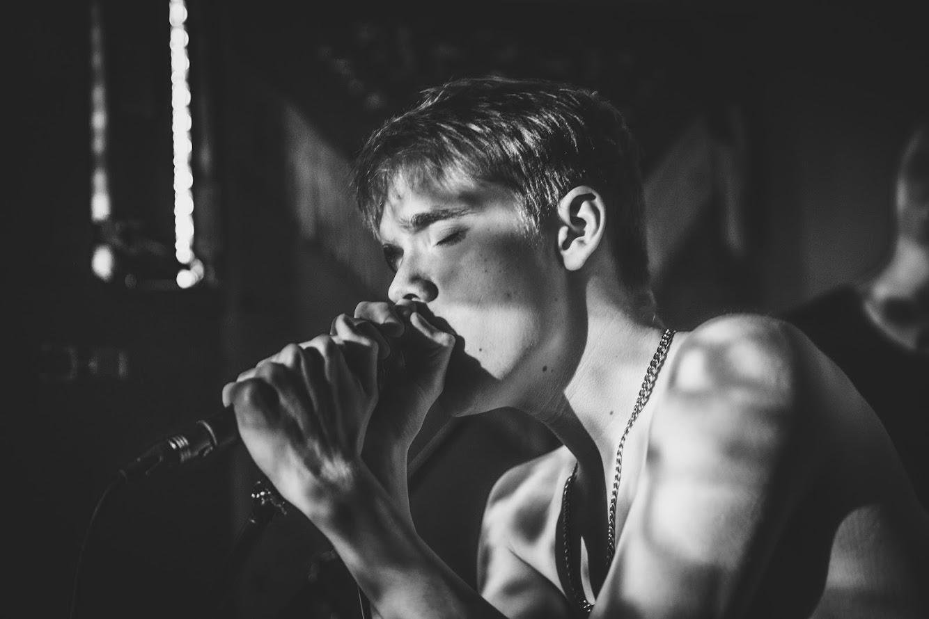 Black and white photo of a person singing into a microphone with closed eyes and a chain necklace.