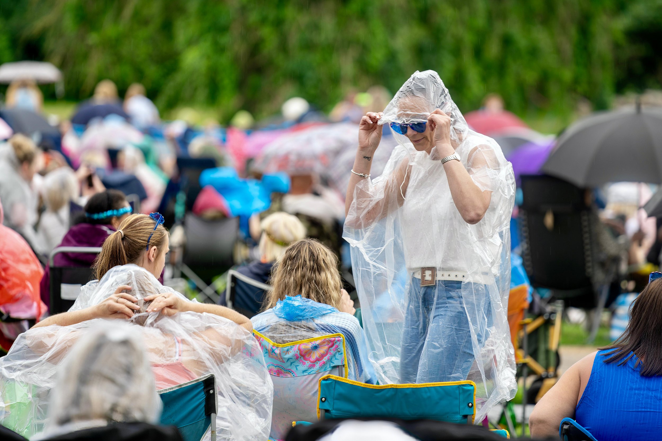 Crowd wearing rain ponchos at an outdoor event, with a person standing adjusting sunglasses.