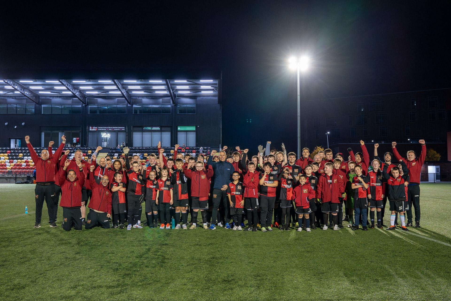 Large group of people, including children, wearing red and black sports attire on a lit soccer field at night, posing together with hands raised in celebration.