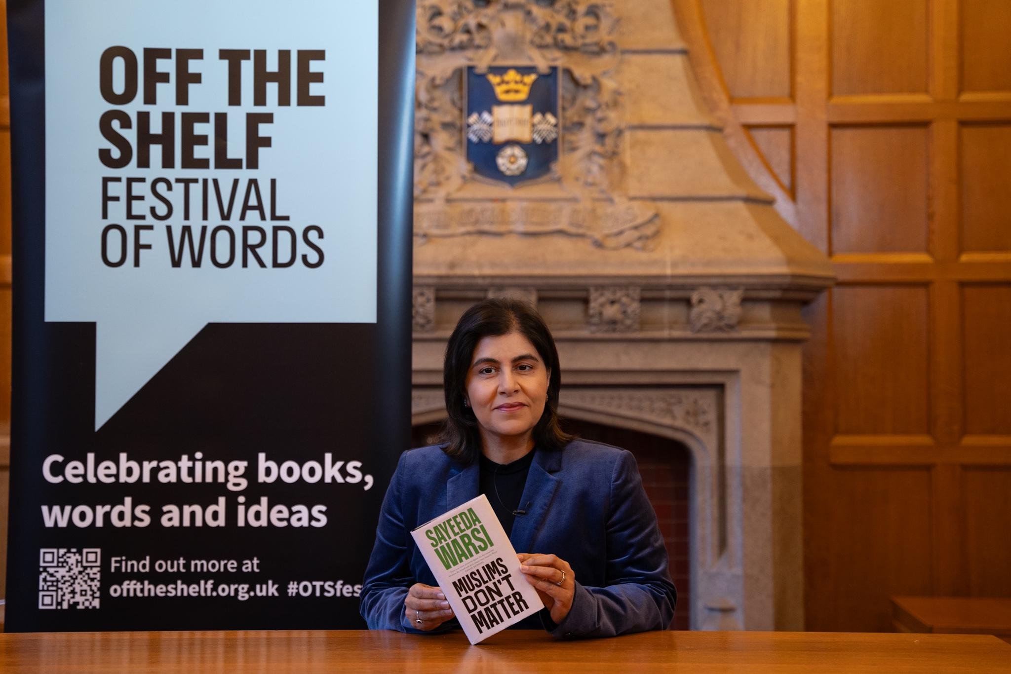 A person holding a book titled 'Muslims Don't Matter' by Sayeeda Warsi, sitting in front of a banner that reads 'Off the Shelf Festival of Words' with 'Celebrating books, words and ideas' written below, set in a room with wooden paneling and a crest