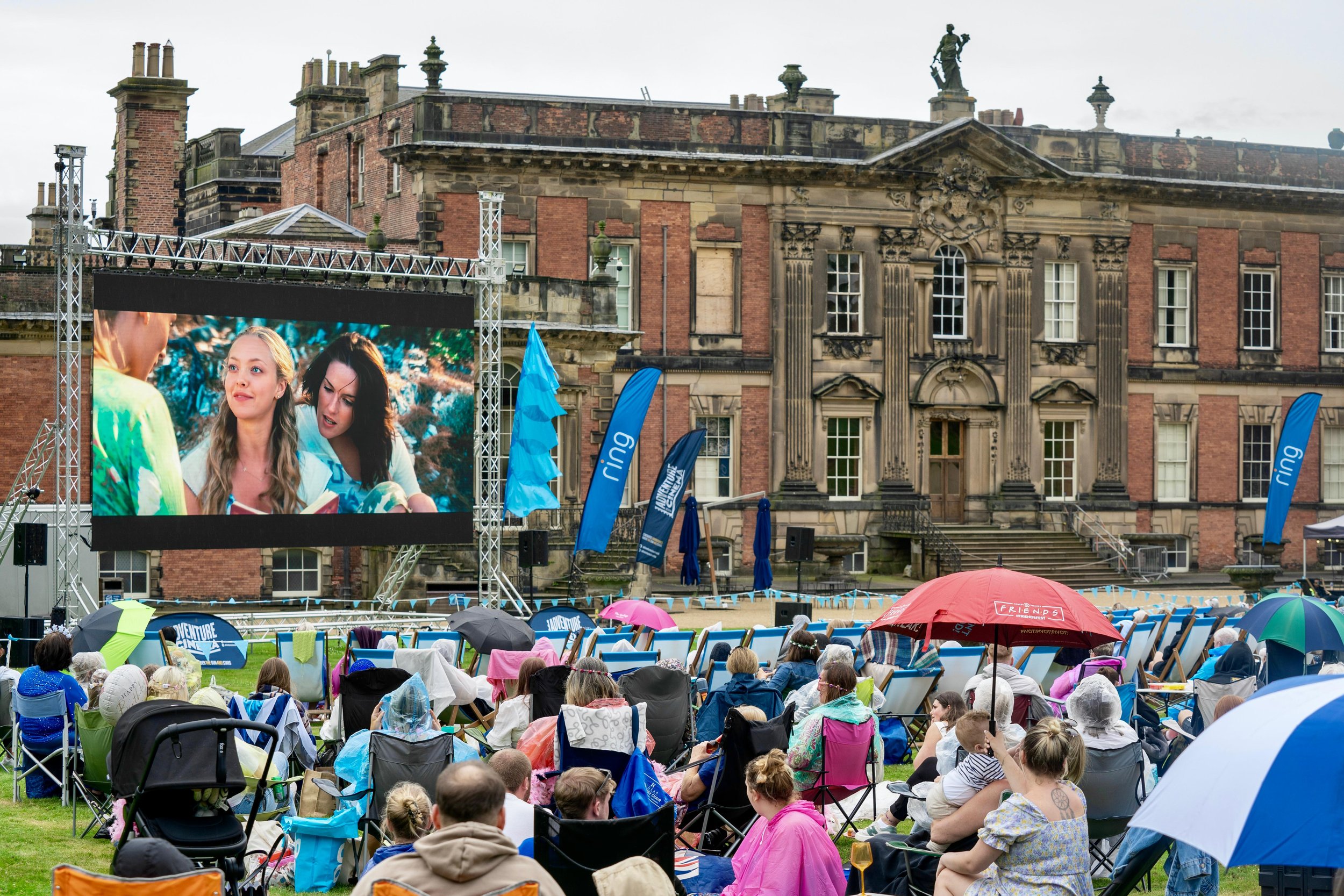 Outdoor movie screening in front of a historic building with an audience seated on chairs and umbrellas in the foreground.