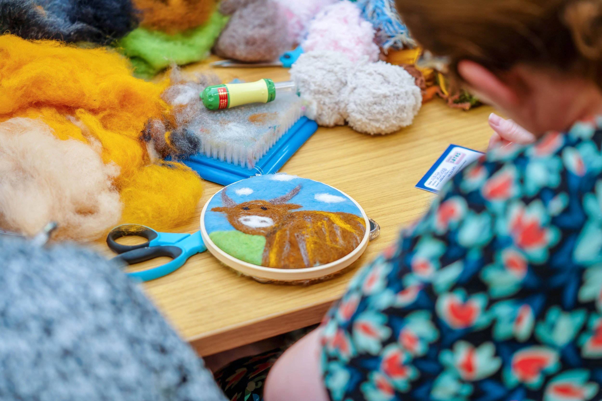 Person creating a felt artwork of a cow, surrounded by colorful wool, scissors, and crafting tools on a wooden table.