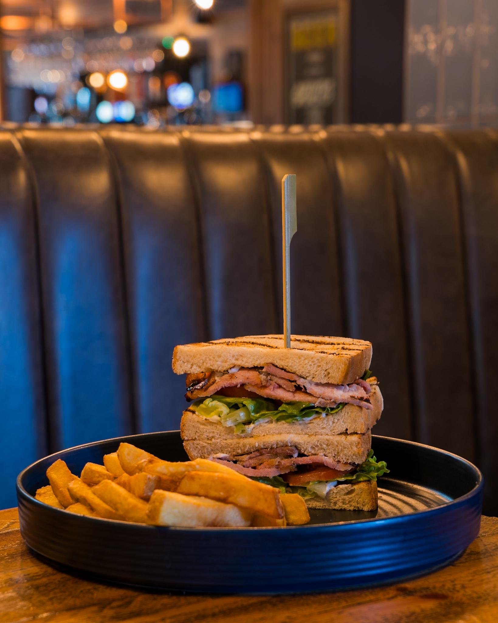 Grilled sandwich with bacon, lettuce, and tomato served with thick-cut fries on a black plate in a restaurant setting.