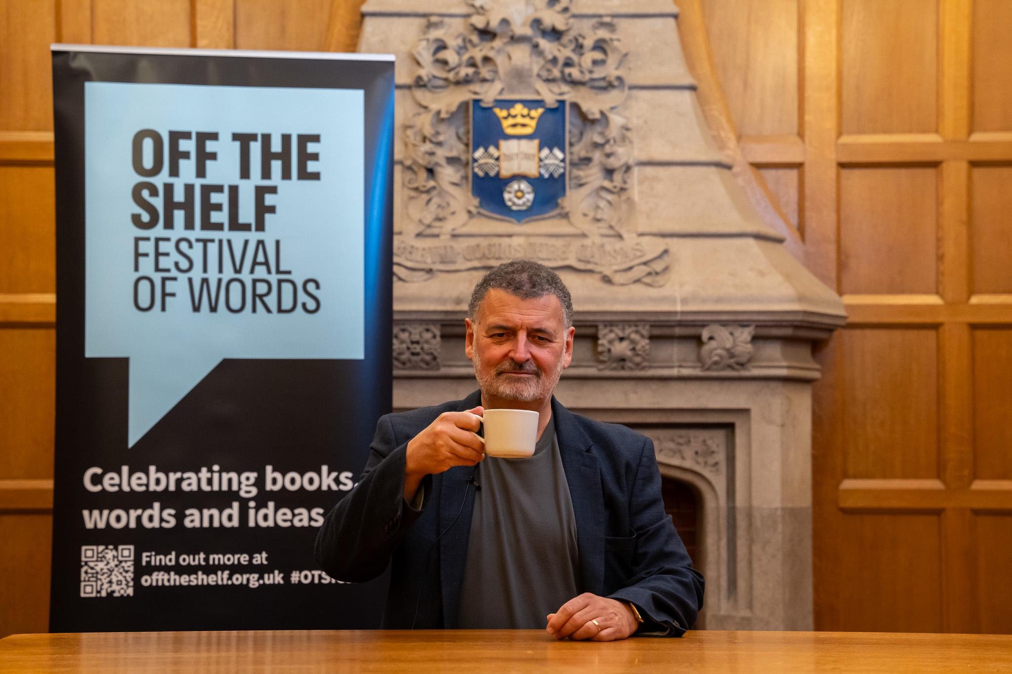 Man holding a cup at a desk with "Off the Shelf Festival of Words" banner.
