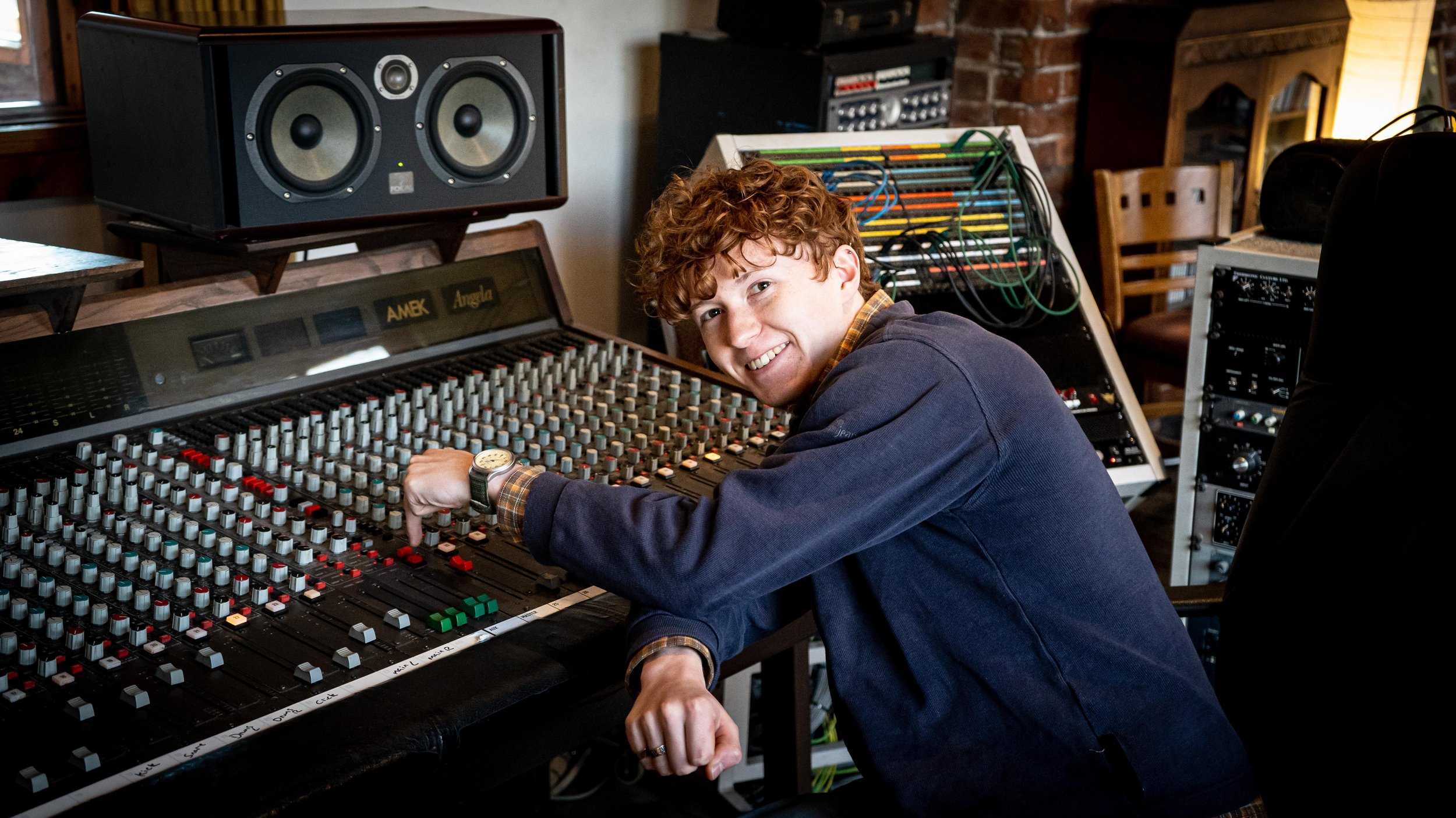 Person adjusting controls on a large audio mixing console in a recording studio, with speakers and cables in the background.