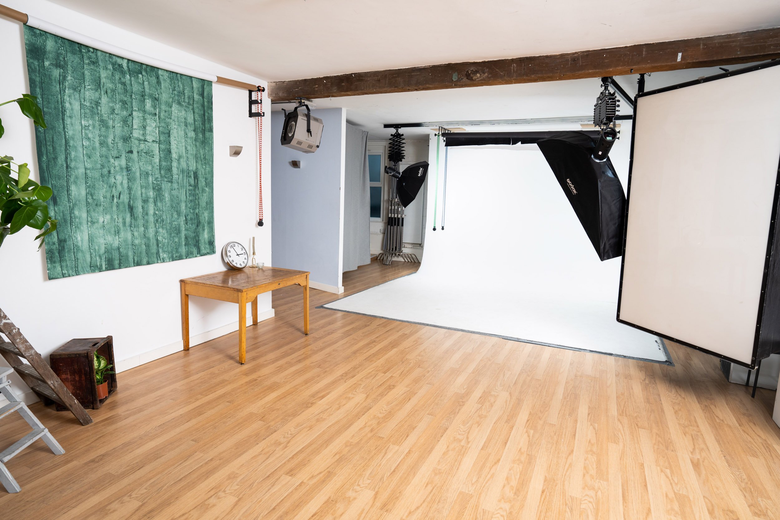 Photo studio interior with lighting equipment, white backdrop, wooden flooring, and a small table with a clock and plant.