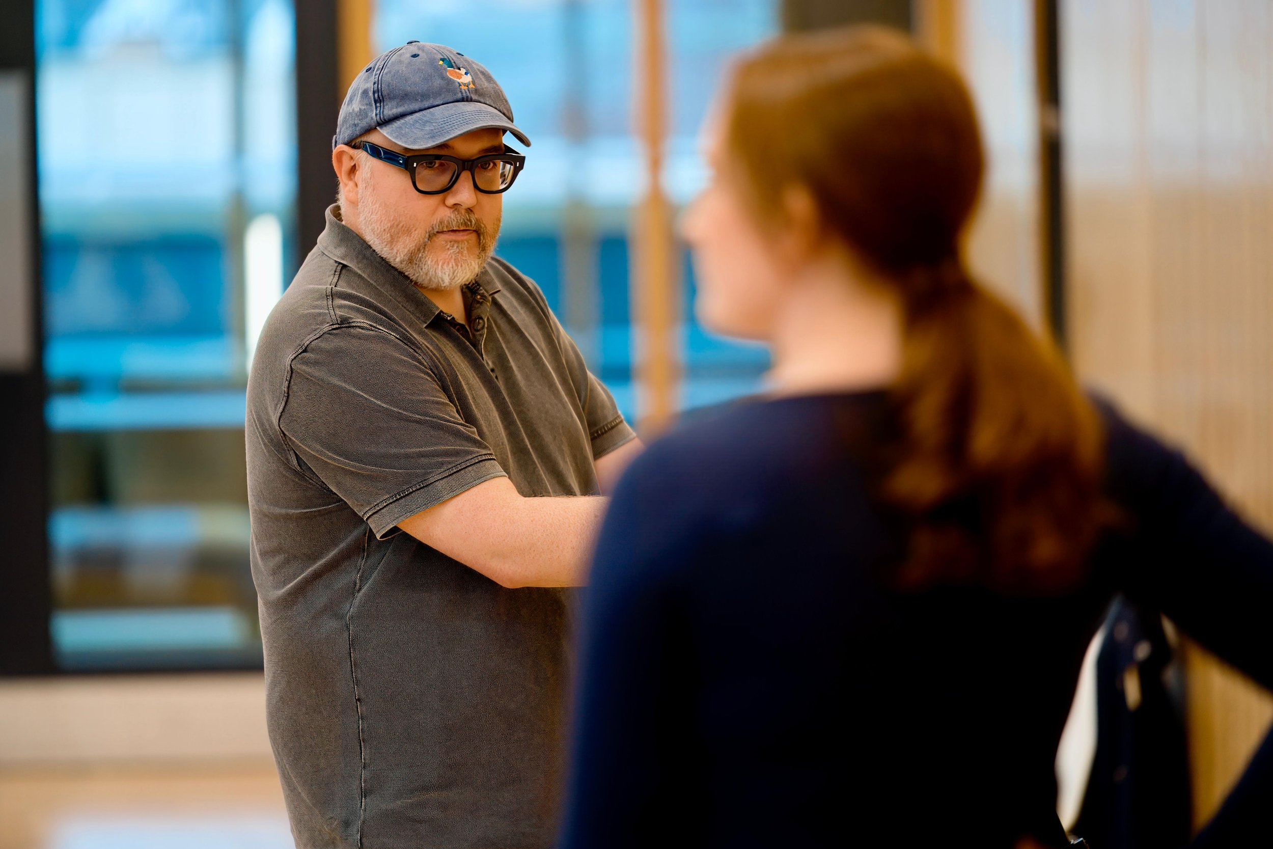Man in glasses and baseball cap looking at a woman in an indoor setting.