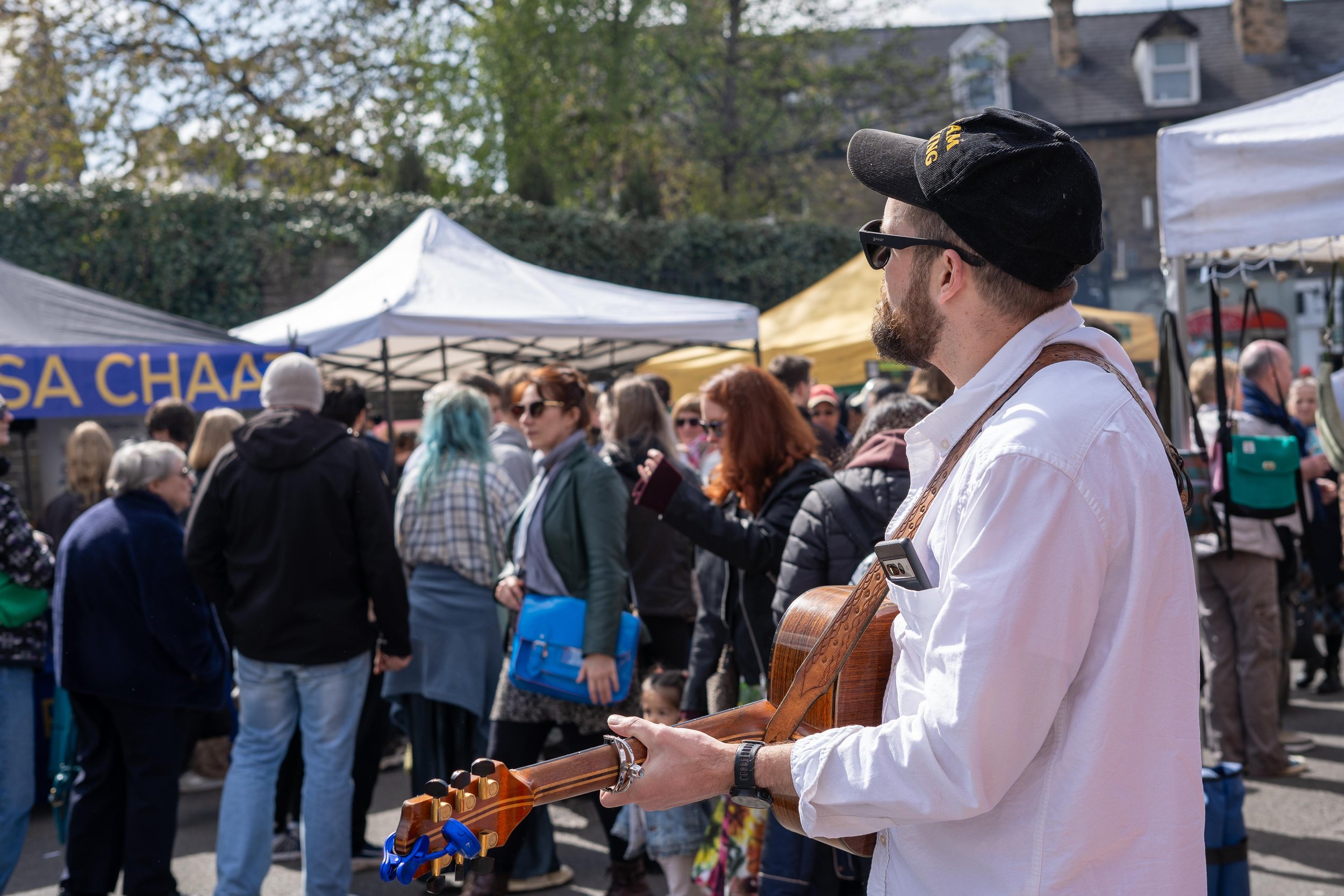 Street performer playing guitar at a crowded outdoor market with tents and people browsing.