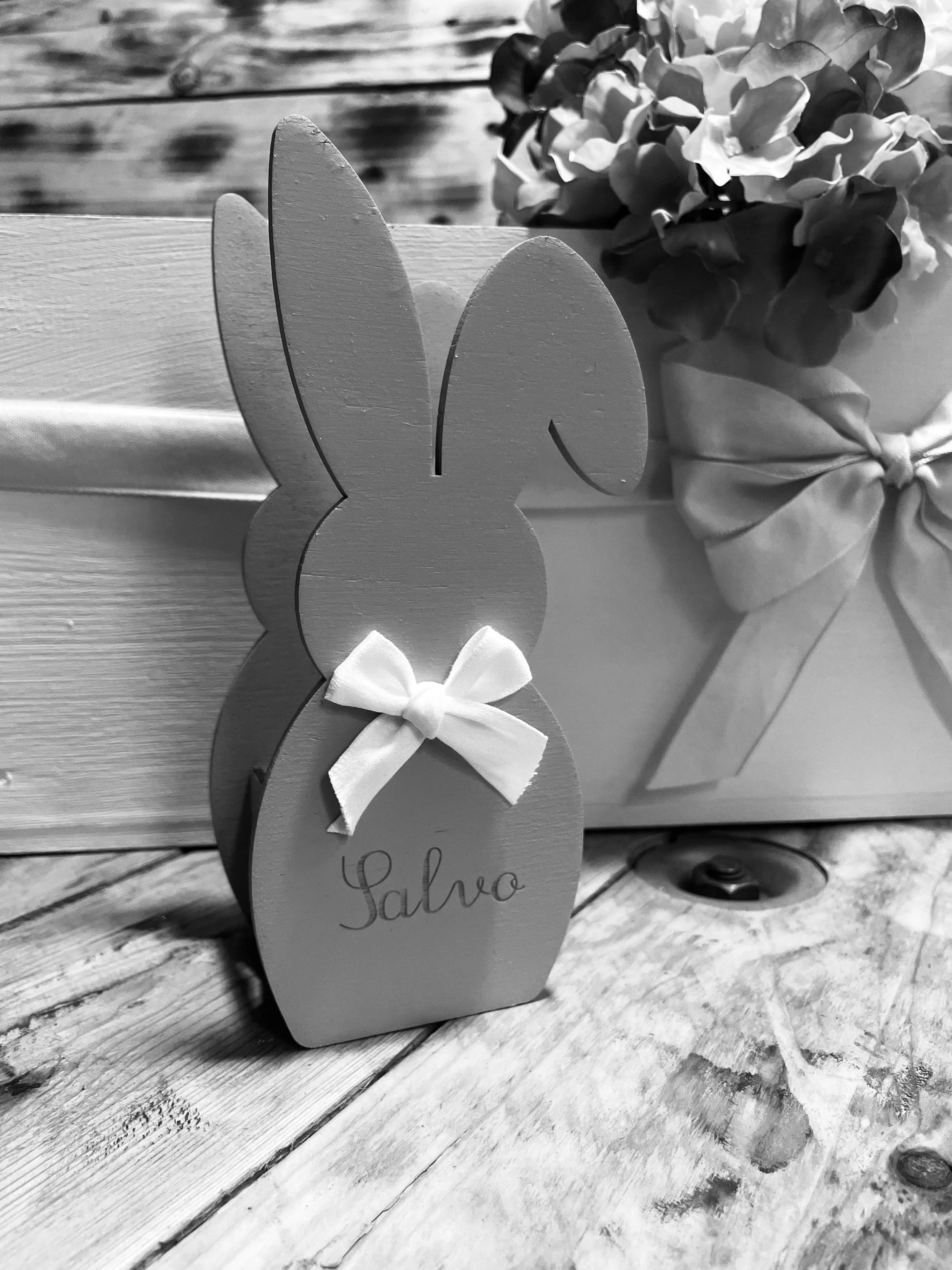 Presina di legno a forma di coniglio decorata con fiocco di stoffa e scritta 'Salvo', posizionata vicino a un vaso con fiori.