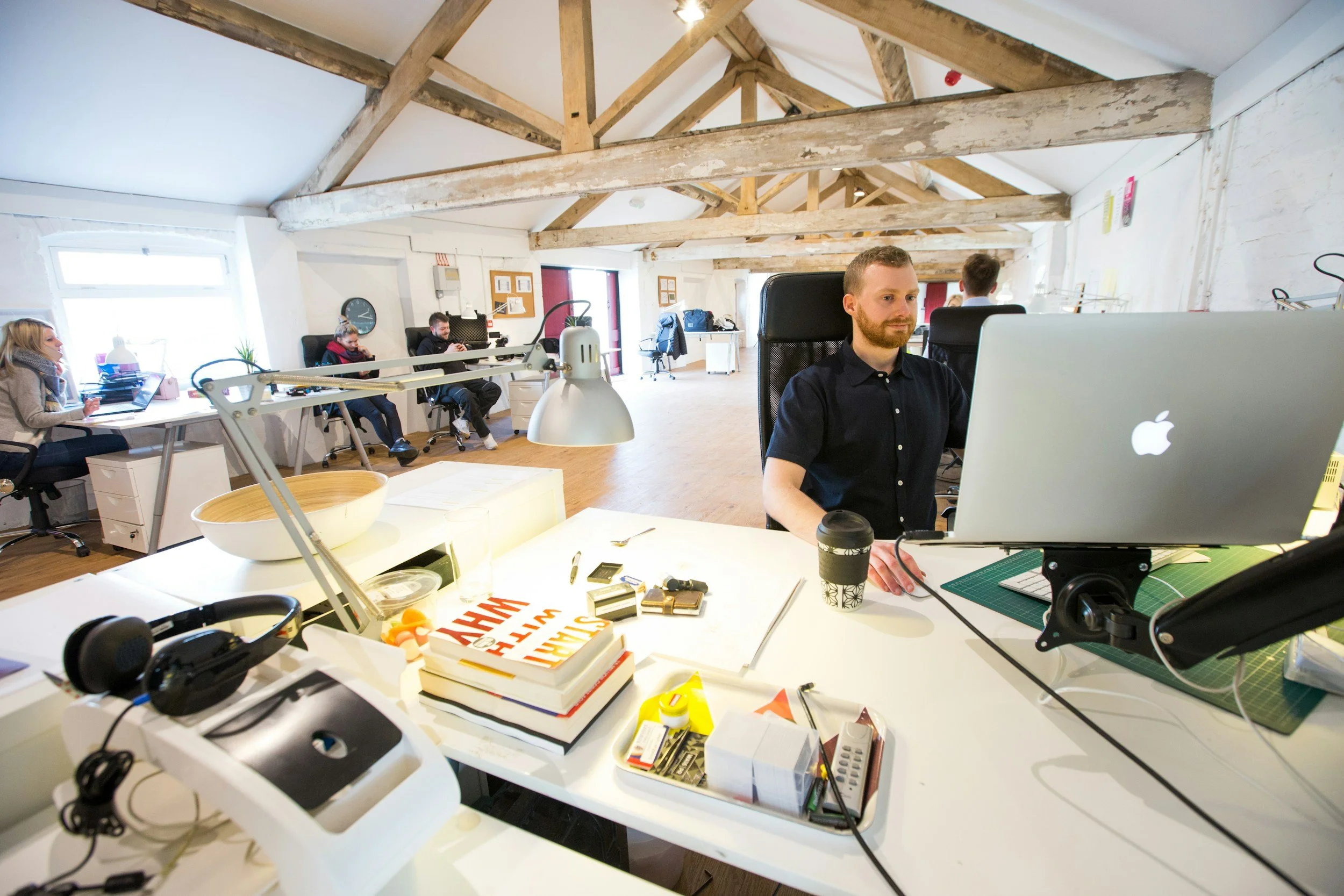 Un bureau moderne avec plusieurs personnes travaillant à leurs ordinateurs, espace lumineux avec poutres en bois apparentes, bureau avec des chaises ergonomiques.
