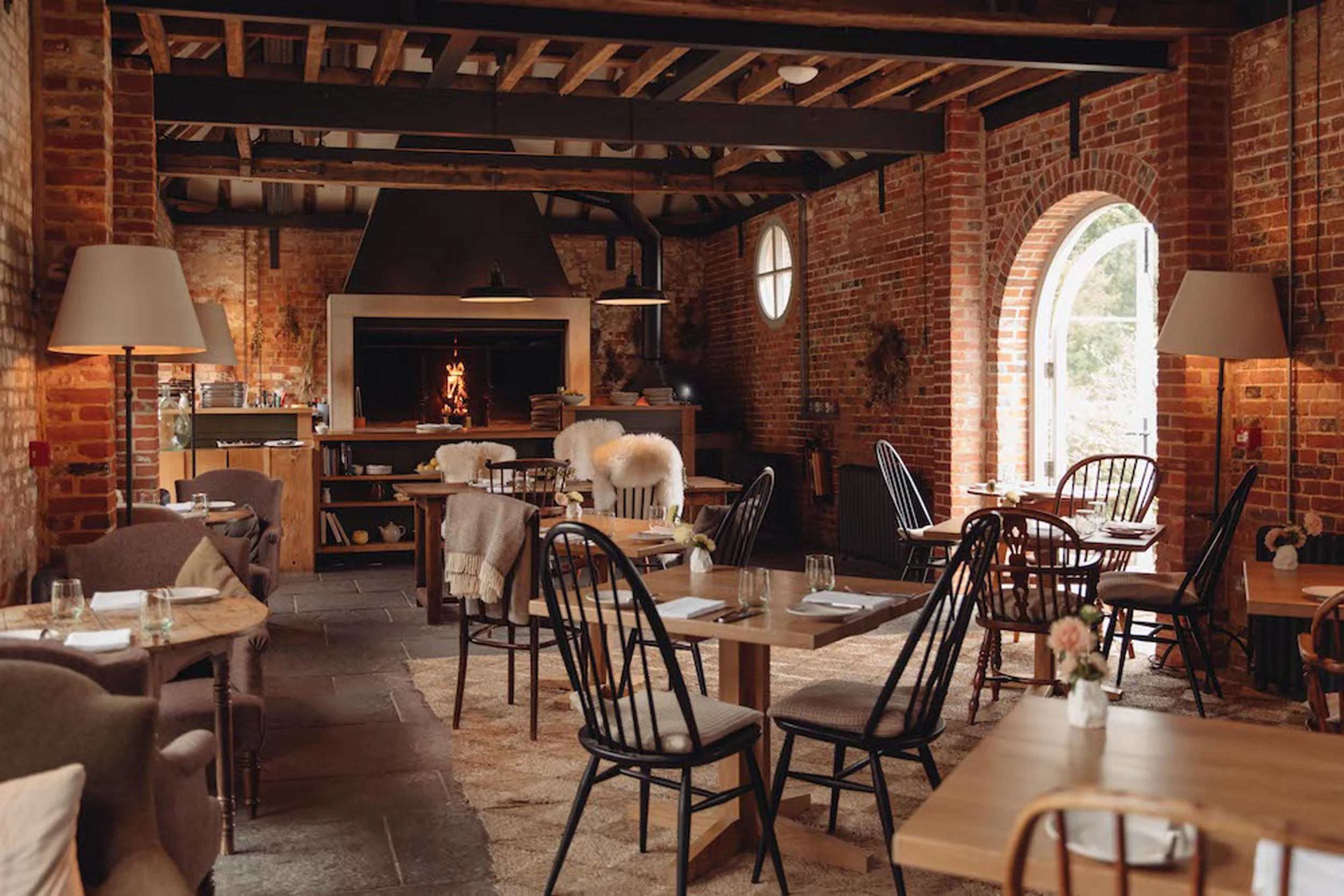Heckfield Place with brick walls, wooden beams, and a cozy fireplace. The dining area features wooden tables and chairs with plush cushions and throws. Natural light enters through large arched windows.