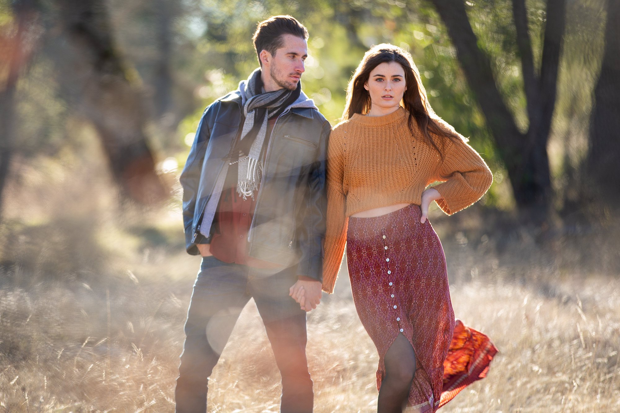 A young couple walking hand in hand through a sunlit field during autumn, holding hands, with the woman wearing a mustard sweater and a long patterned skirt, and the man dressed in a jacket and scarf.