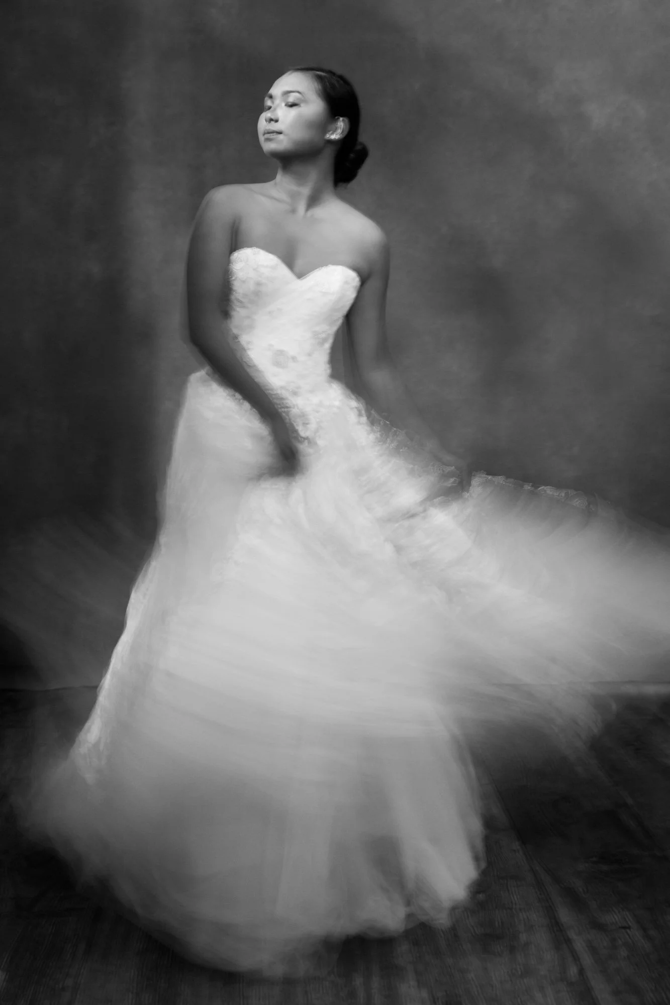 A woman in a wedding dress standing on a wooden floor. The photo is black and white, with a blurred motion effect on the skirt.