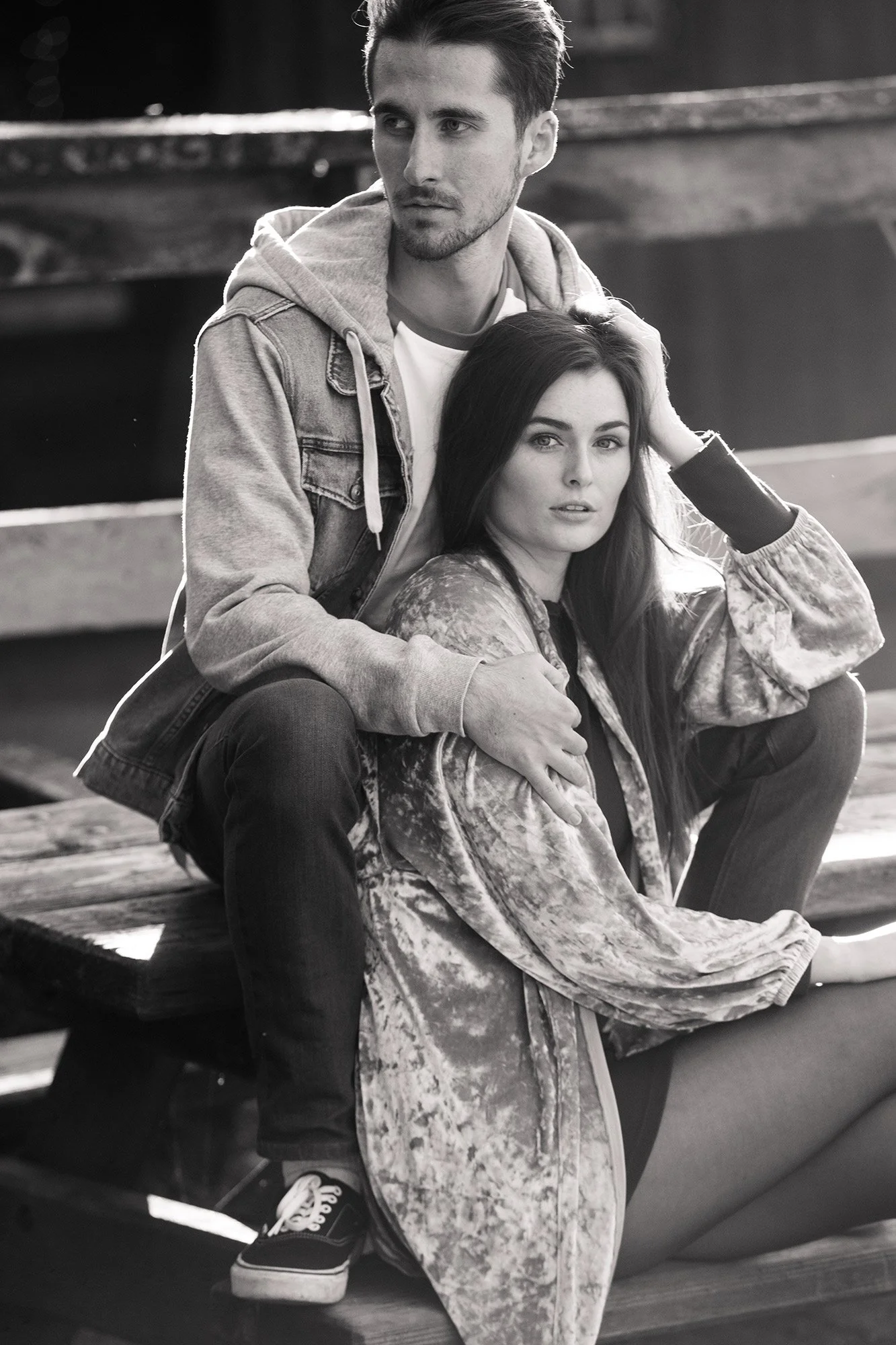 A black and white photograph of a man and woman sitting on a wooden bench outdoors. The man has short hair, facial hair, and wears a denim jacket with a hoodie. The woman has long dark hair and wears a patterned jacket. They are looking at the camera