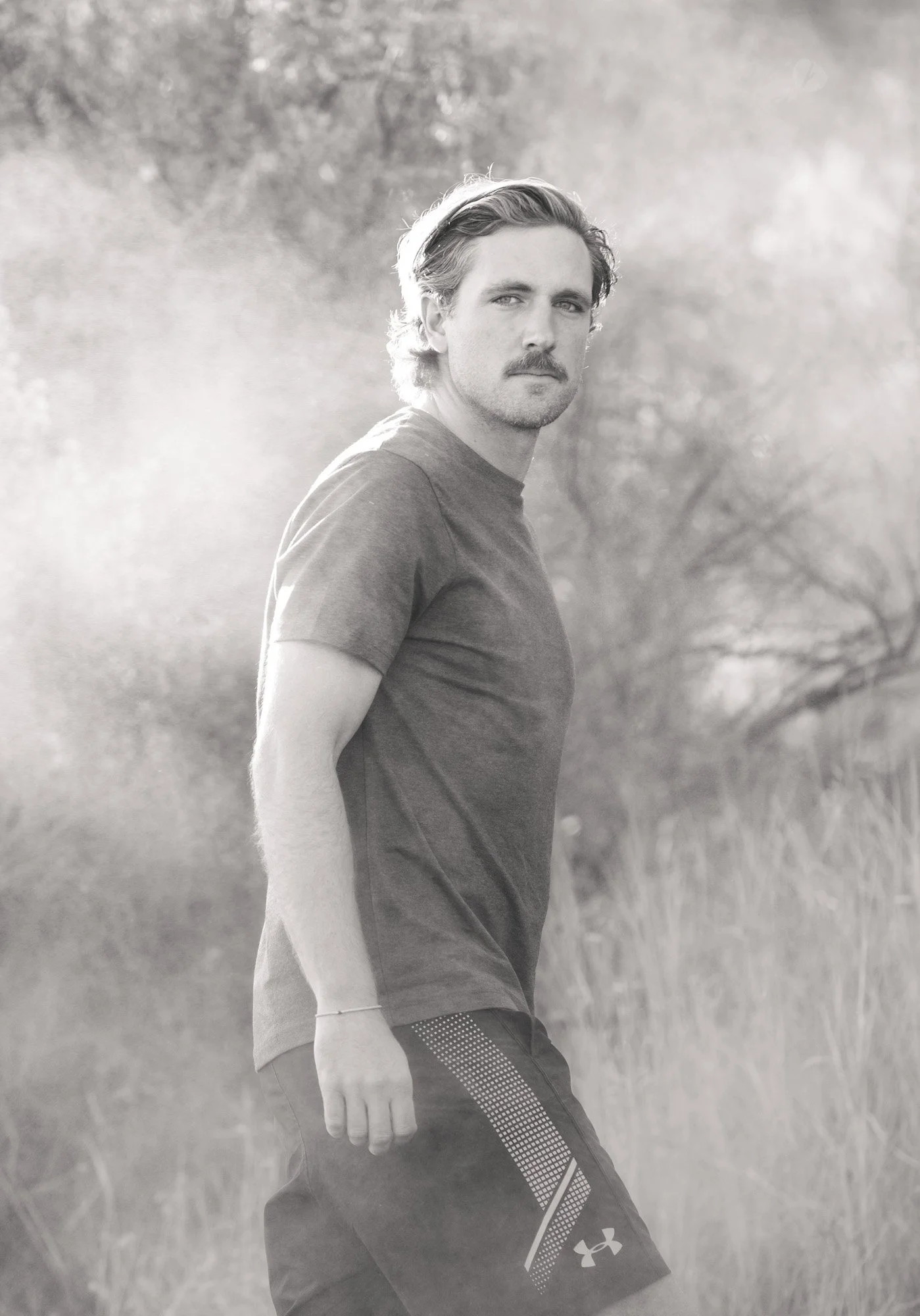 A black and white photo of a man with light-colored hair and a mustache, standing outdoors in a natural setting with trees and grass, wearing a short-sleeved shirt and shorts, looking at the camera.