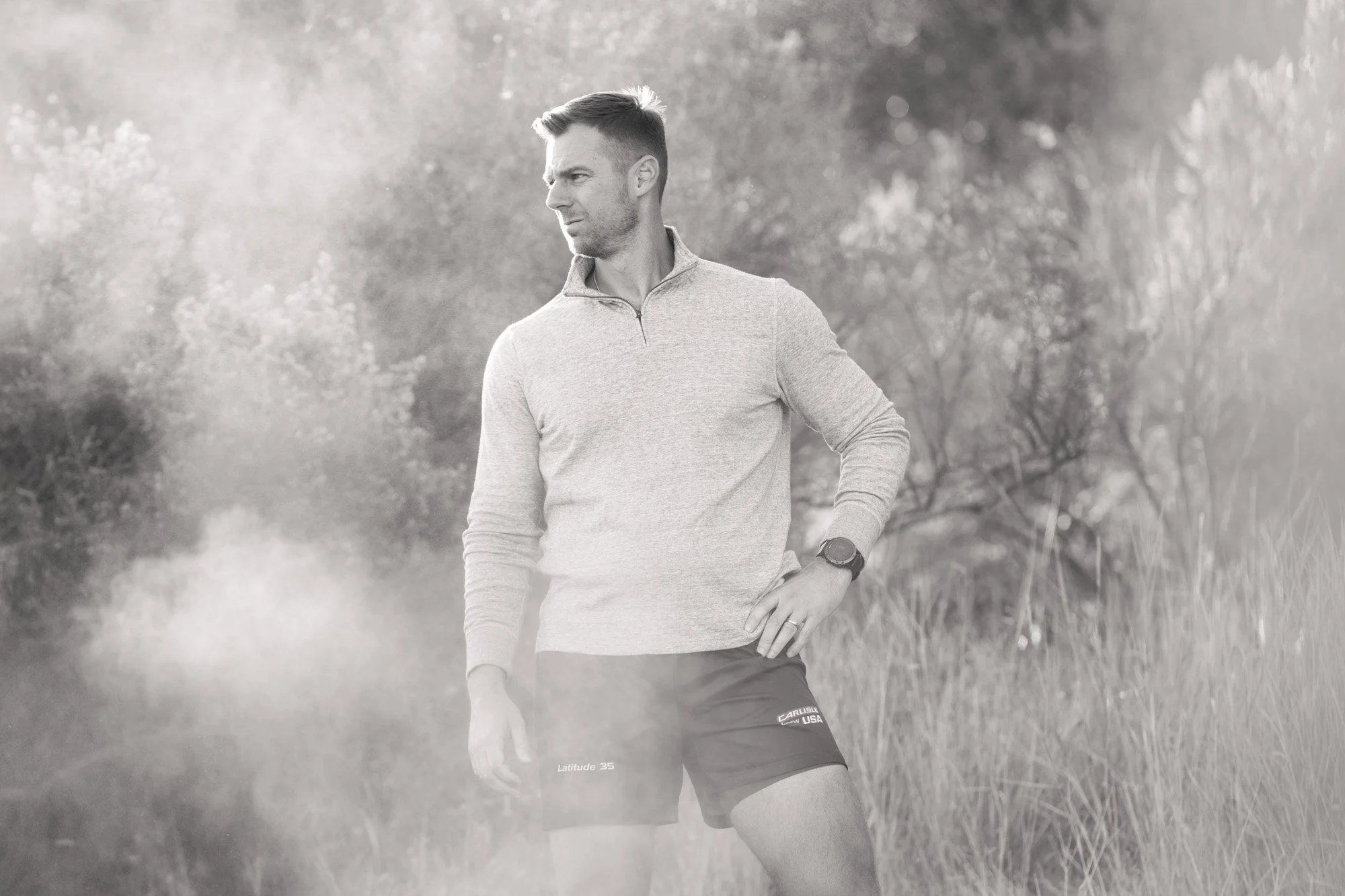 A man in athletic clothing standing outdoors in a grassy area, looking to the side with a serious expression, with trees and bushes in the background.